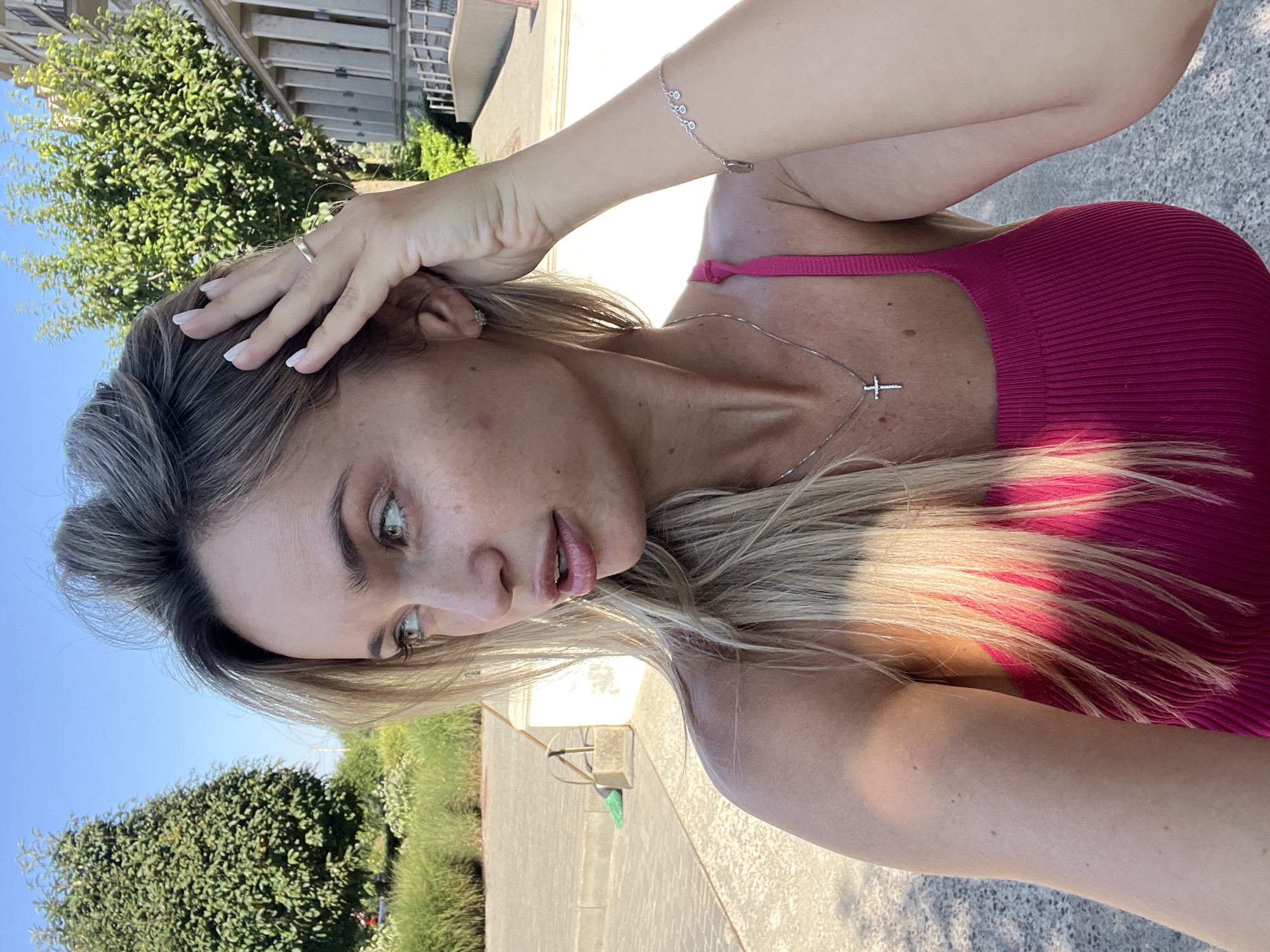A young woman with blonde hair, wearing a pink ribbed tank top and a silver necklace with a cross pendant, poses for a selfie outdoors on a sunny day. Her left hand is raised to her head, and she wears a silver ring on her index finger and a silver bracelet on her left wrist. The background shows a clear blue sky, lush green trees, and a building with columns. The ground appears to be paved with interlocking stones, and in the middle ground, a green broom and dustpan are visible next to a white structure. The scene is set in Fontanka, Ukraine.