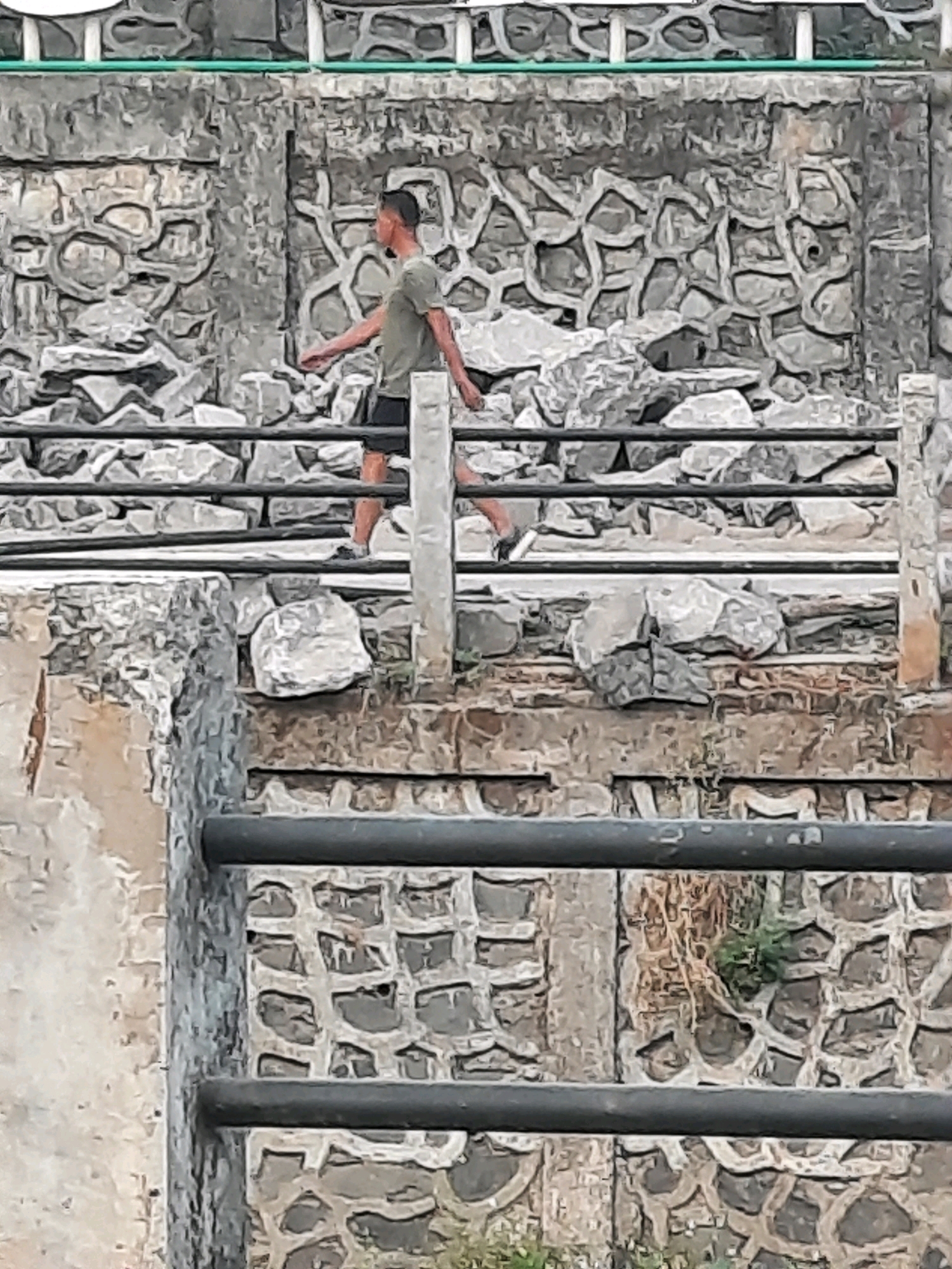 The image shows a man walking along a raised walkway in East Jakarta, Indonesia. The walkway is made of concrete and has black metal railings. Behind the walkway is a stone wall with a decorative pattern. The man is wearing a green t-shirt and black shorts. He is walking to the left of the frame. The environment appears to be outdoors, with some vegetation visible in the background. The lighting suggests it is daytime.