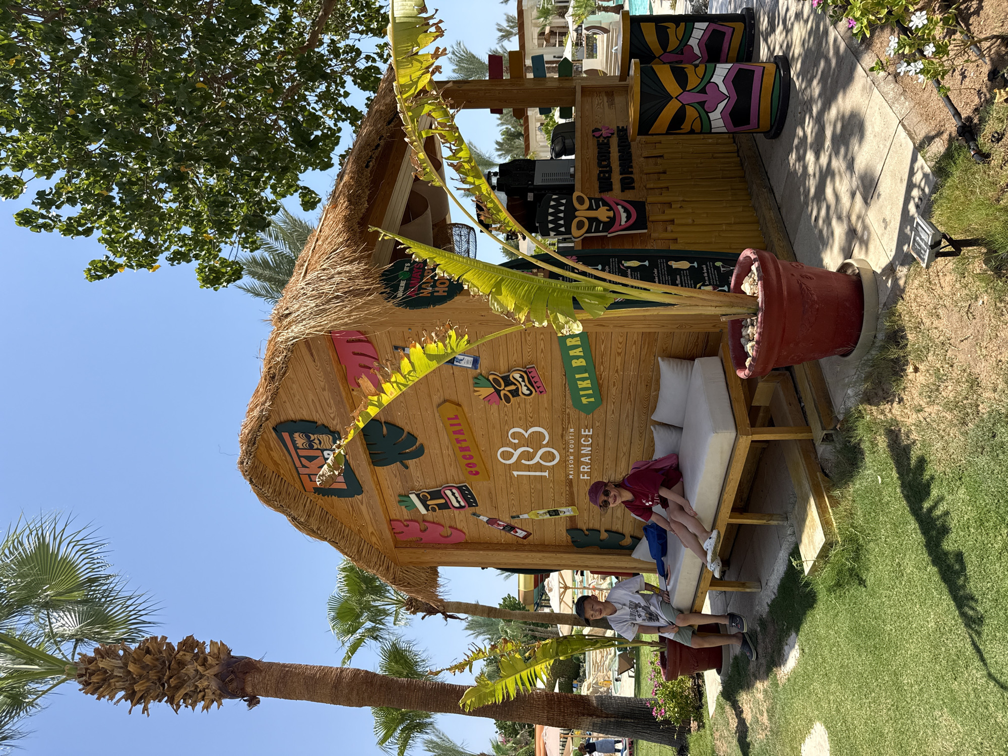 The image depicts a vibrant outdoor scene at a tropical resort, likely in Sharm El Sheikh, Egypt, given the context. At the center is a charming, rustic tiki bar constructed from wood with a thatched roof. The bar is adorned with colorful Polynesian-themed masks and graphics, including a sign that reads "TIKI BAR" and another that says "WELCOME TO PARADISE". A young boy and girl are seated on a cushioned bench in front of the bar, appearing relaxed and enjoying the sunny atmosphere. The boy wears a grey t-shirt and shorts, while the girl has on a maroon t-shirt, shorts, and a cap. Lush green grass surrounds the bar, with palm trees and other tropical foliage adding to the exotic ambiance. In the background, glimpses of swimming pools and resort buildings suggest a holiday destination. The overall impression is one of a cheerful, vacation-oriented setting.