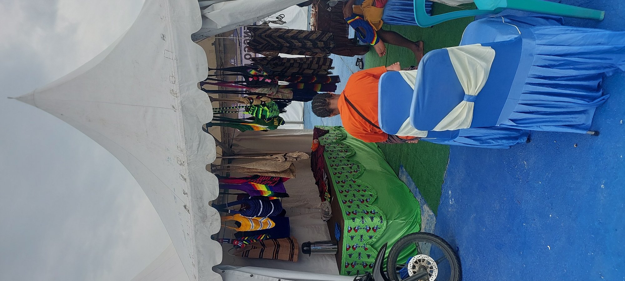 In Asei Besar, Indonesia, a white tent structure stands open, displaying various handmade items for sale. The tent is supported by a central pole and has a peaked roof. Underneath the tent, colorful woven bags hang from a horizontal bar. A long table covered with a green fabric featuring an abstract pattern is set up, holding a metal thermos and other items. To the left, the front wheel of a motorcycle is visible. In the foreground, two blue chairs are draped with blue fabric skirts and adorned with cream-colored sashes tied into bows. A person wearing an orange t-shirt is seated on one of these chairs, facing away from the camera. Another person's legs are visible on the right side of the image, sitting on a blue draped chair. The ground is covered with blue carpeting.