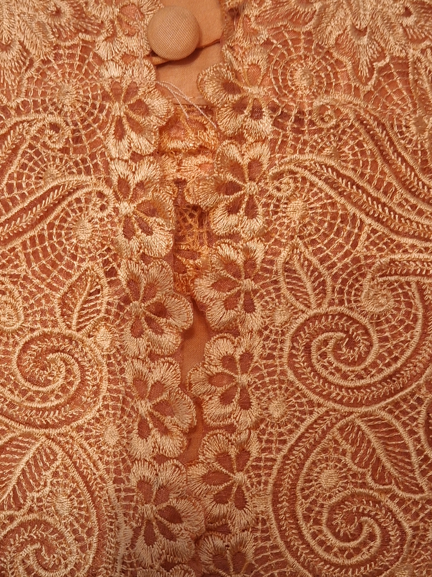 The image displays a close-up view of intricate orange lace fabric. The fabric features a repeating pattern of floral motifs and swirling, curvilinear designs, creating a dense and textured appearance. Some areas of the lace are slightly sheer, revealing the underlying fabric. A portion of a rounded, ribbed orange button is visible at the top center of the image. The lighting is even, highlighting the detailed embroidery and the subtle variations in the fabric's color. There are no discernible elements or context in the image to suggest a location in Lumajang, Indonesia, or any urban environment. The image is purely a detailed shot of textile.