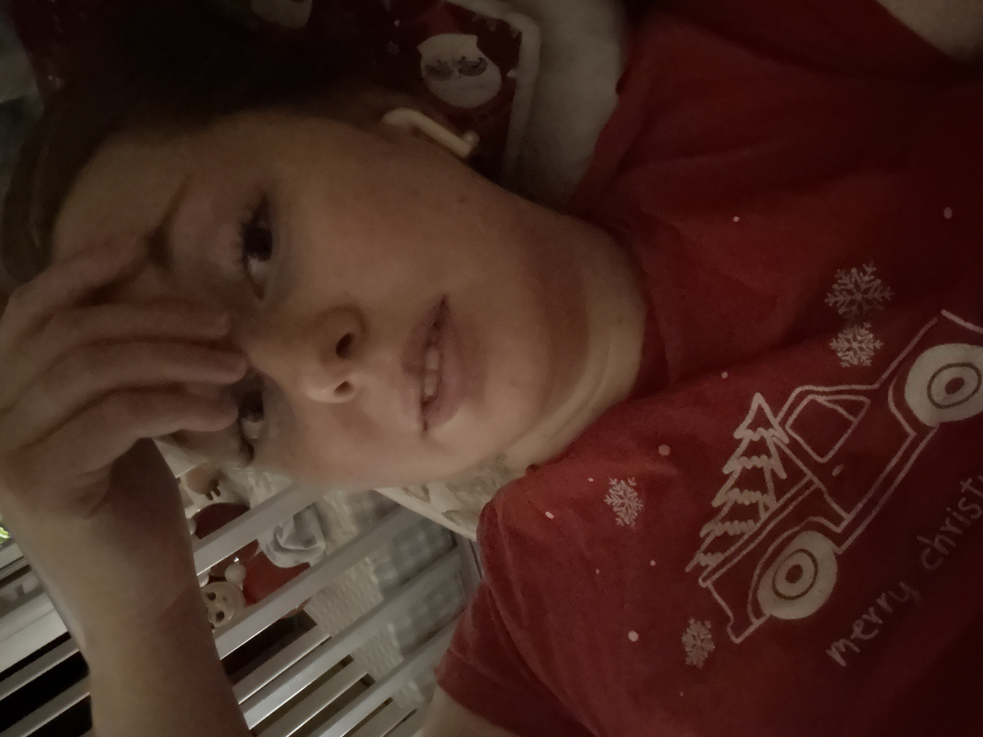 A person, likely an adult female, is shown lying down, looking towards the camera. They are wearing a red short-sleeved t-shirt with a white graphic design. One hand is raised to their forehead, covering part of their eye, suggesting an expression of tiredness, thought, or slight discomfort. The person's mouth is slightly open.
The graphic on the t-shirt features a white outline of a vintage-style pickup truck with a Christmas tree in its bed. Several white snowflakes are scattered around the truck. Partially visible white text below the truck reads "merry christ", likely part of "merry christmas".
The background is dimly lit, suggesting an indoor setting, possibly at night or in low light conditions. Behind the person's head, there's a white pillow or blanket, and a hint of red fabric with a Santa Claus pattern can be seen. To the left and bottom of the frame, a series of vertical white slats are visible, indicative of a crib or a bed frame. These slats suggest the person might be in a nursery or a bedroom, possibly leaning into a crib.
The overall scene conveys a relaxed, yet potentially pensive or tired mood, set during the Christmas season.