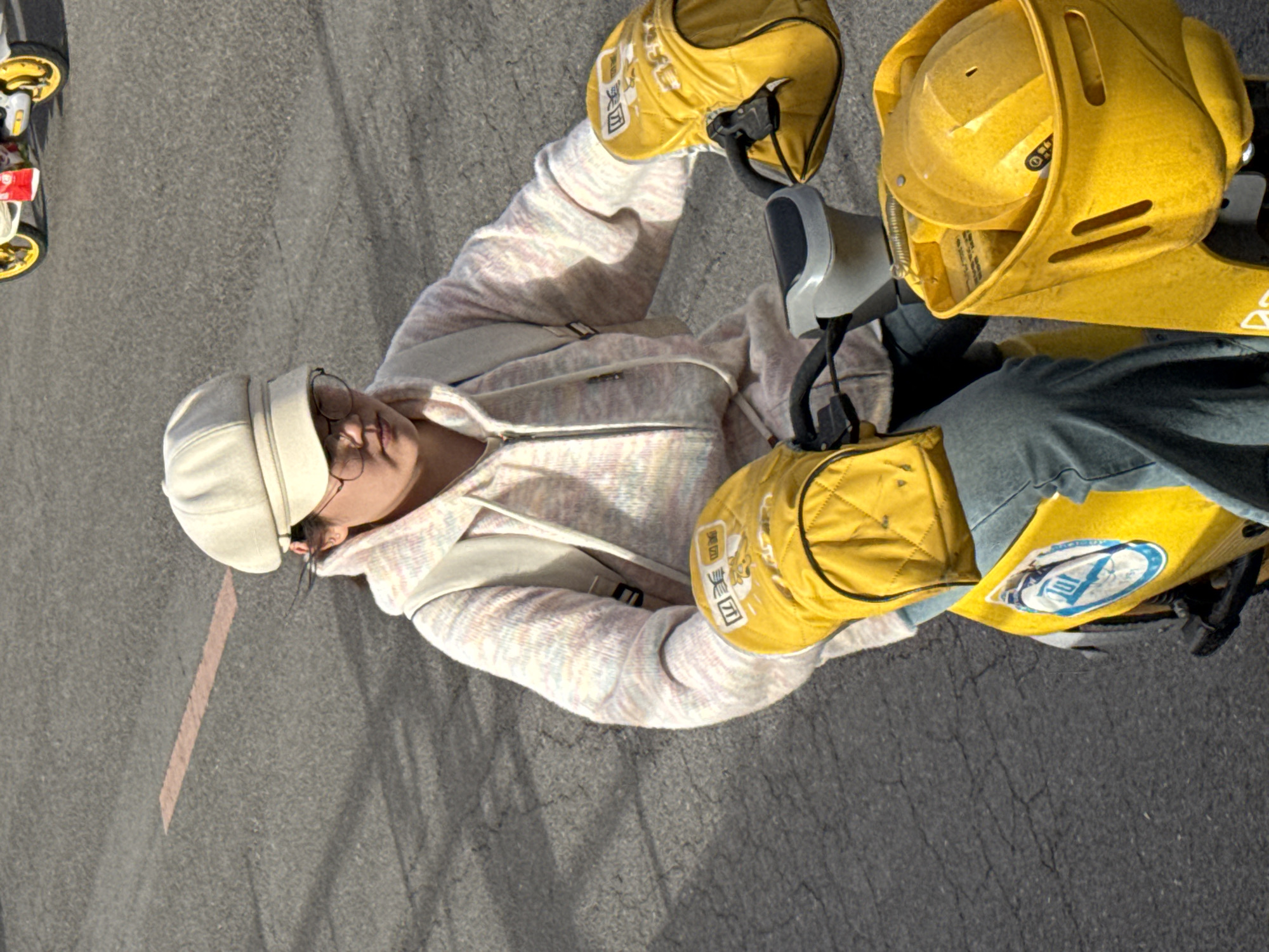 A high-angle shot shows a person wearing a white hat and glasses sitting on a yellow electric scooter. The scooter has yellow padded containers attached to the handlebars and back. The person wears a pastel-colored zippered hoodie and jeans. Another small go-kart-like vehicle is visible in the upper left corner. The setting appears to be on a paved road in Zhongtun, China.