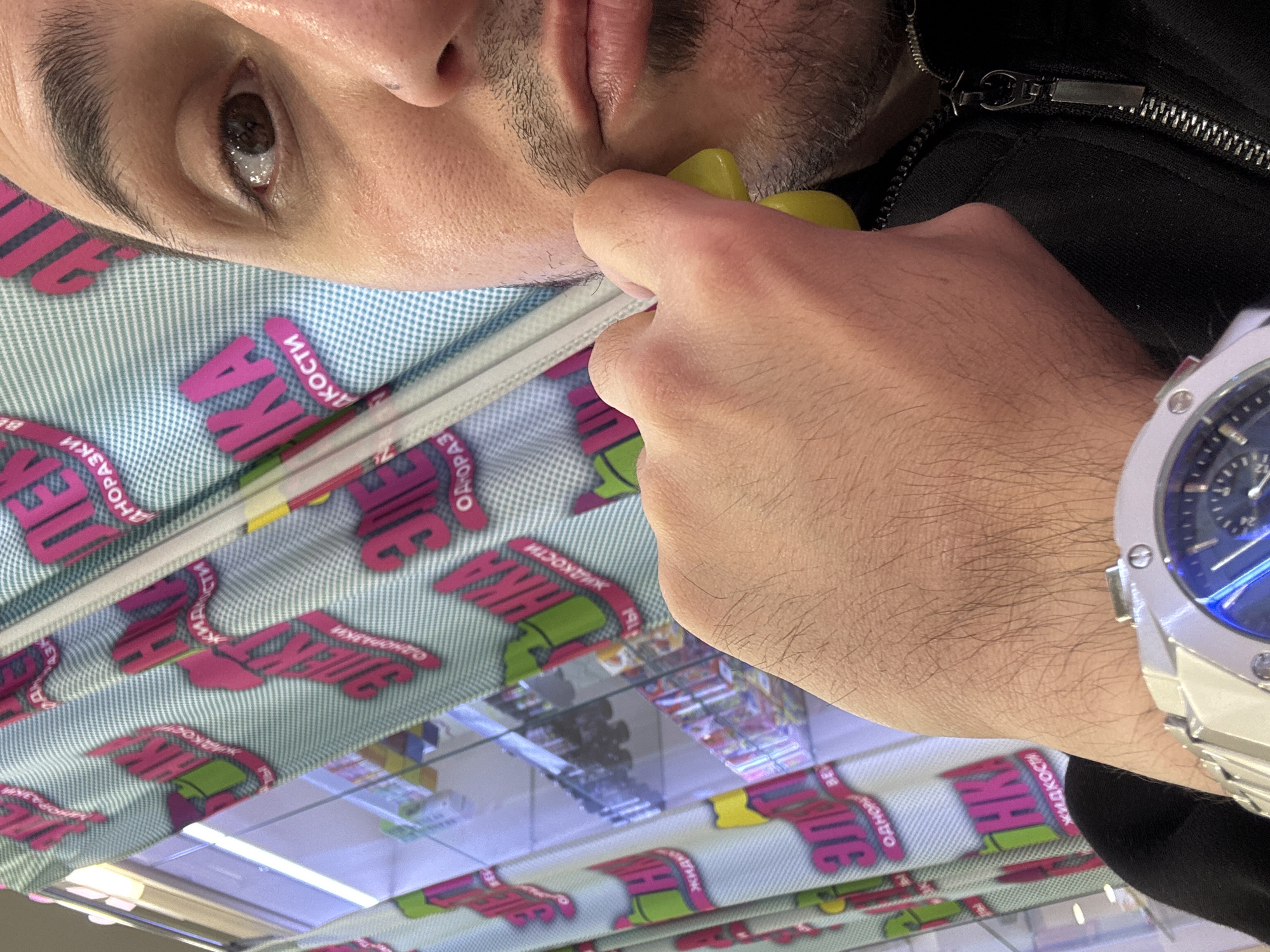 A selfie taken from a low angle shows the face of a man with brown eyes, dark eyebrows, and stubble on his chin. He is wearing a black zip-up jacket and a silver watch with a blue face. His hand is near his face, holding a yellow object. The background is a patterned surface with text in Russian. The text reads "ЖИДКОСТИ ОДНОРАЗКИ", which translates to "Disposable Liquids". The patterns and reflections suggest the photo was taken inside a store selling vaping products in Saint Petersburg, Russia.