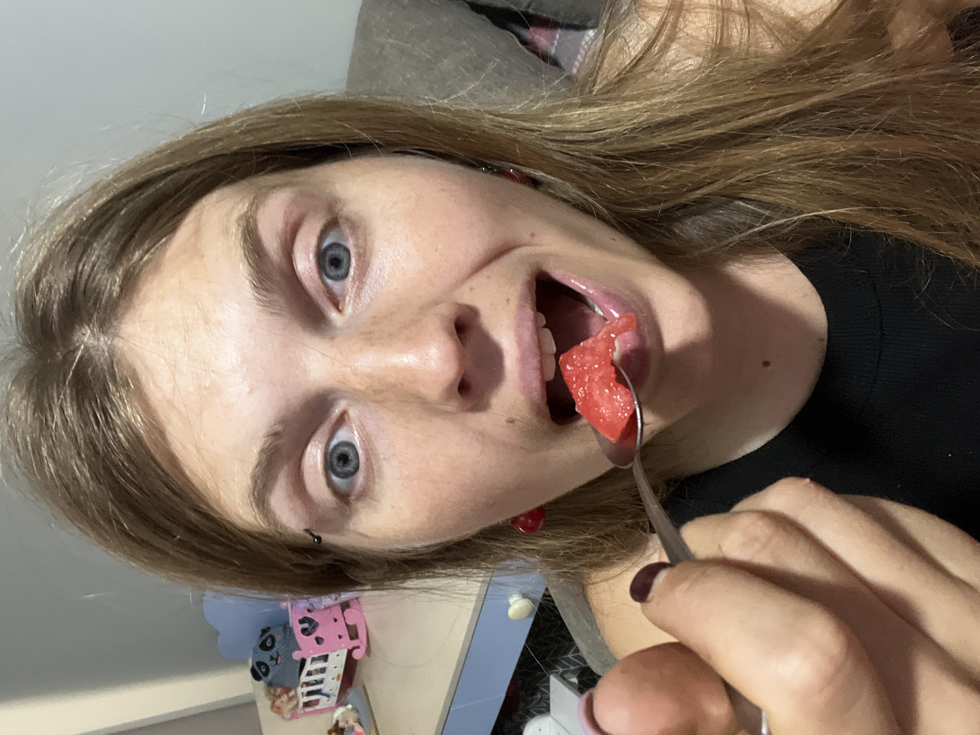 A woman with blonde hair and blue eyes is captured in a close-up, selfie-style shot, holding a spoon with a piece of watermelon towards her open mouth. She is wearing a black top and red cherry-shaped earrings. A piercing is visible on her left eyebrow. In the blurred background, a child's bed with a panda toy and a light blue heart-shaped headboard can be seen, suggesting a domestic setting, possibly in a bedroom or nursery. The lighting appears to be artificial. The location in Сеніцкі сельскі Савет, Belarus, is noted.