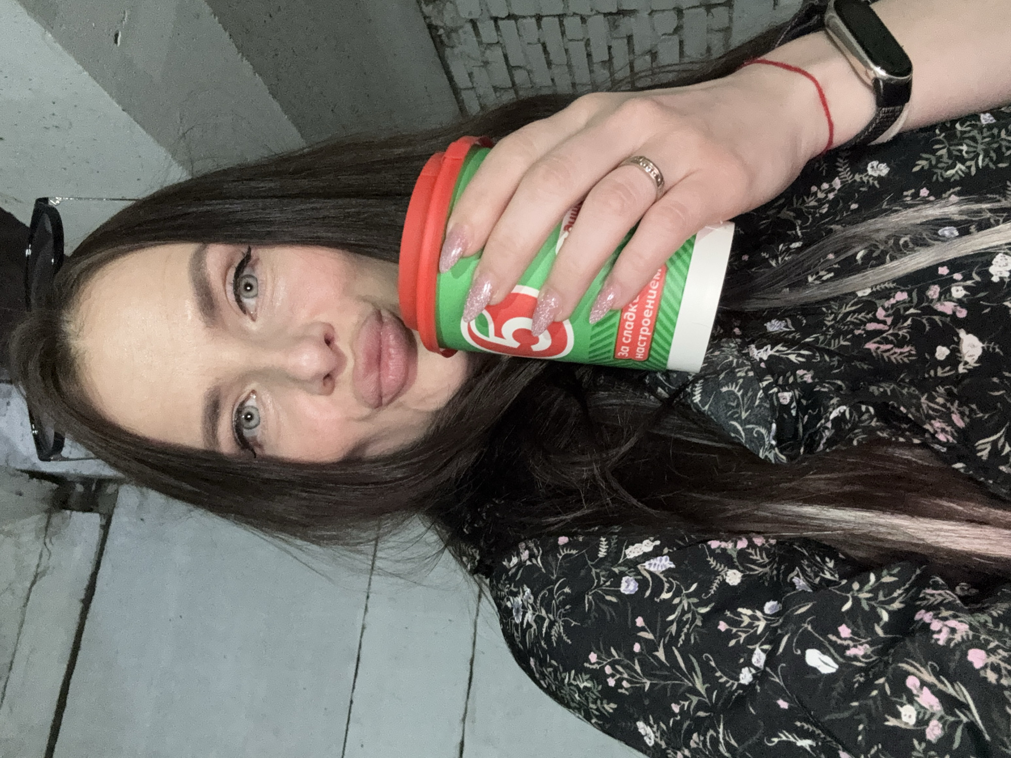 A woman is shown in a selfie-style photo, holding a disposable coffee cup with a red lid and green body with Russian text. She is making a pouting expression. She has dark hair with a light streak, fair skin, and a floral print dark-colored dress. She is wearing a ring and a fitness tracker on her wrist. The background is a wall of concrete blocks, possibly a parking garage, in Chaikovsky, Russia.