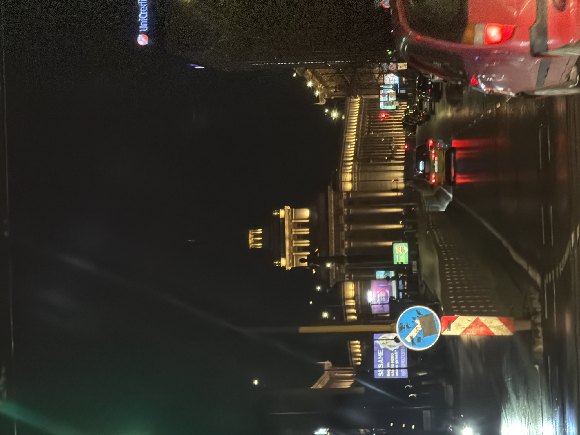 The image depicts a night scene on a street in Sofia, Bulgaria. Illuminated by streetlights and car headlights, the cityscape includes prominent buildings with classical architectural features. Traffic is present, indicated by car tail lights and the presence of a red vehicle in the upper right corner. Road signage, including a blue directional sign and a red and white striped barrier, are visible in the foreground. Billboards and advertisements light up the background, notably a "Unicredit" sign and another one that reads "SESAME." The road surface appears wet, likely due to rain.