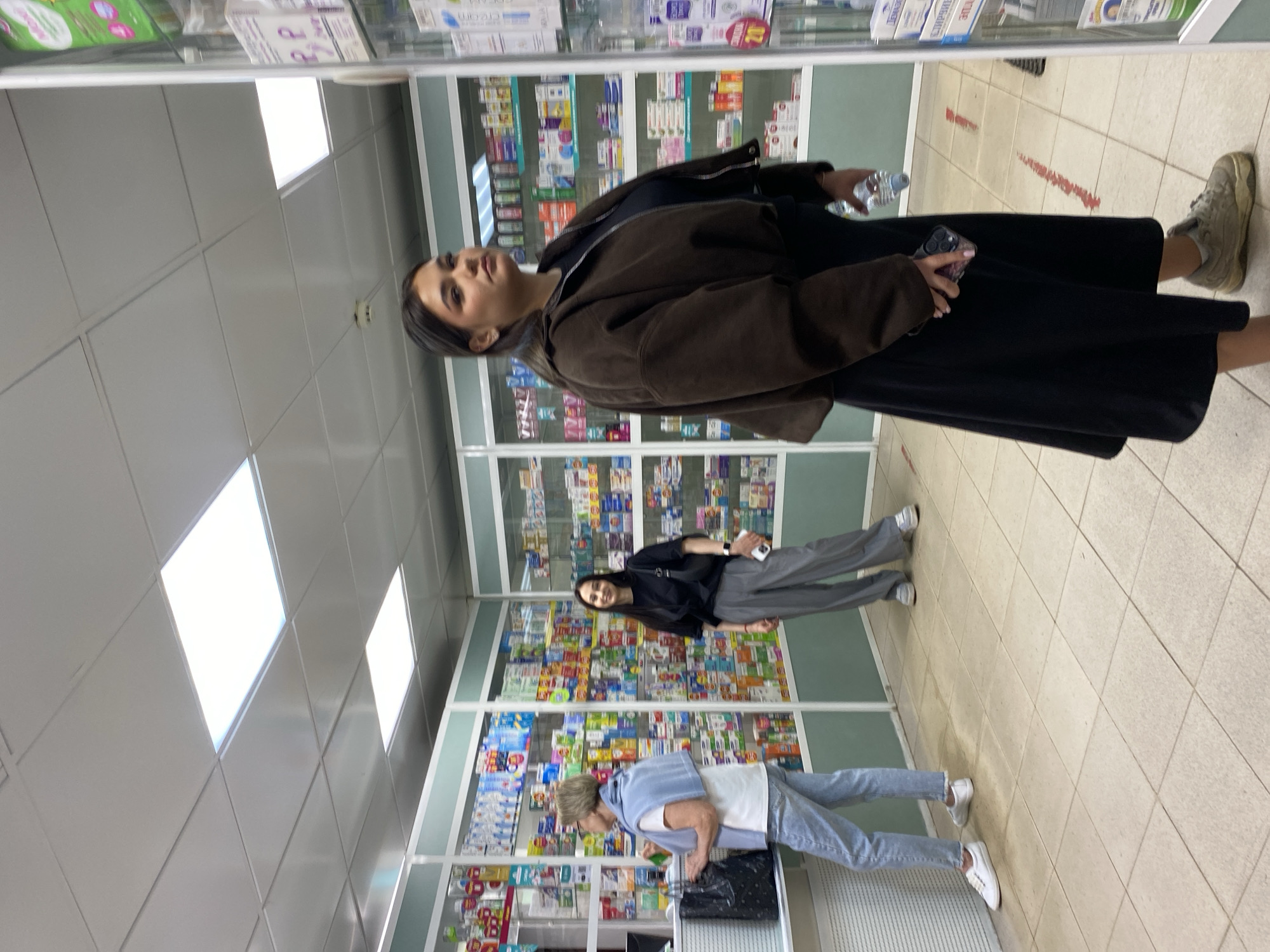 The image shows an interior view of a pharmacy, likely in Chaikovsky, Russia, judging by the medication packaging. Three people are visible. A woman wearing a black dress, brown jacket, and sneakers is standing in the foreground, holding a phone and a water bottle. Further back, another woman in a black top and grey pants stands next to a medication display. An older woman in a light blue top and jeans is bent over a bag at the back. The background is dominated by shelves stocked with various medications. The floor is tiled. The ceiling is tiled and lit with fluorescent panels.