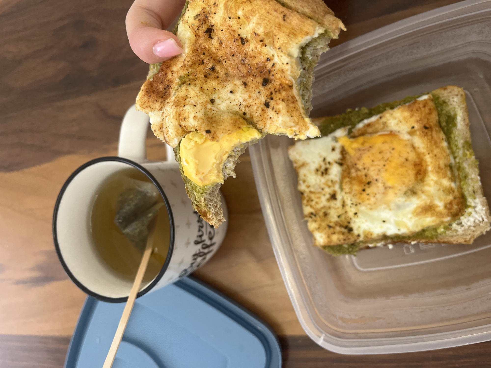 The image captures a top-down view of a casual meal set on a wooden surface. A person's hand, displaying light pink nail polish, holds a partially eaten piece of food. This food appears to be a thick slice of bread or focaccia, topped with a fried egg, revealing a bright yellow yolk where a bite has been taken. A green spread, possibly pesto, is visible between the bread and the egg. The egg and bread are seasoned with dark specks, likely pepper or other spices.
To the right, another similar piece of food, featuring a fried egg on green-spread bread, rests inside a clear plastic food container. To the left, a white ceramic mug with a black rim contains a light yellowish-green liquid, indicative of tea, with a triangular tea bag submerged. A thin wooden stir stick is placed within the mug. The mug features partial visible text in a decorative font, reading "but first,". A light blue plastic lid is partially visible beneath the mug. The scene suggests a relaxed meal, likely breakfast or brunch.