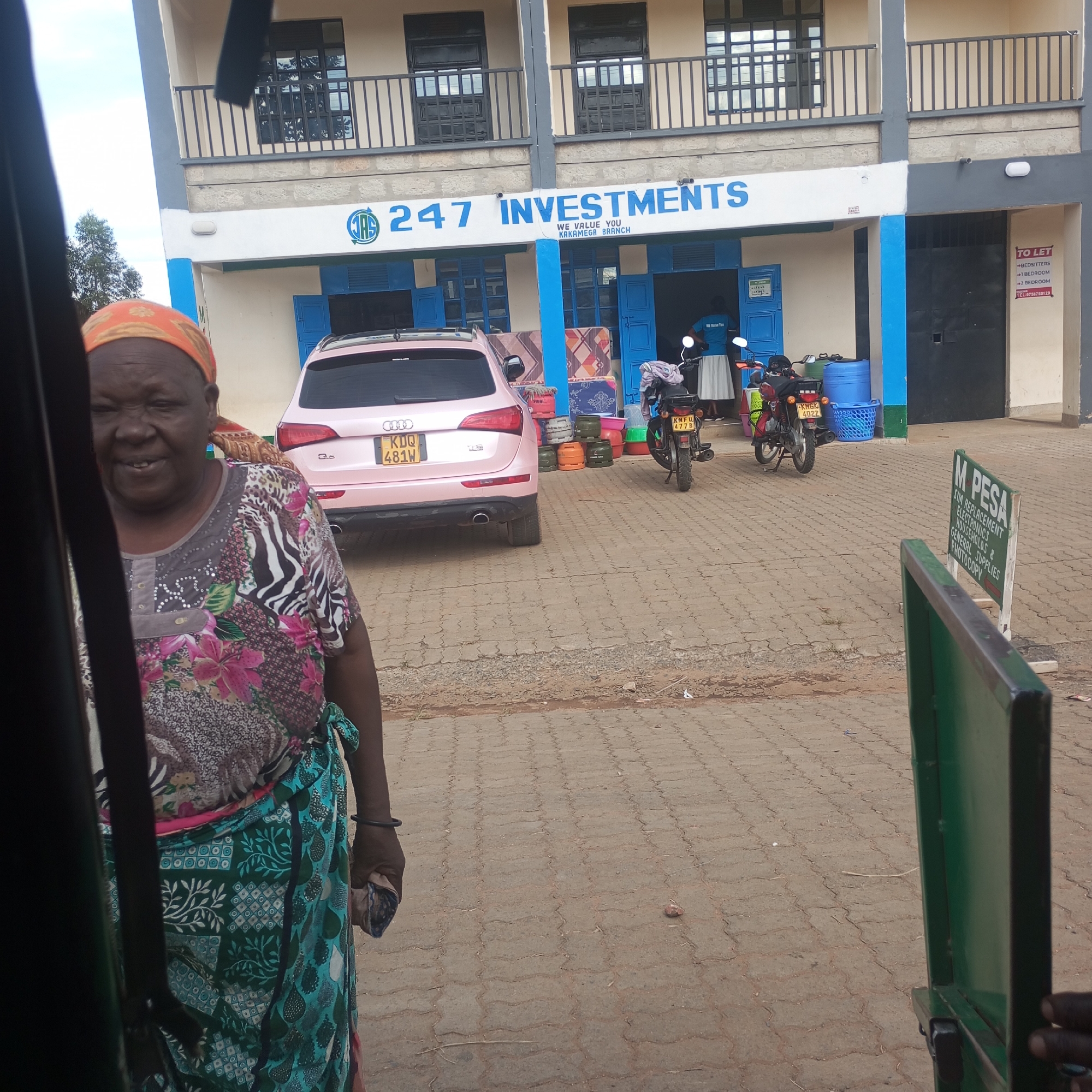 The image shows a view from inside a vehicle of a woman standing near a street in Sheywe ward, Kenya. The woman wears a colorful floral top, a green and blue patterned skirt, and an orange headscarf. Behind her, a pink Audi SUV with license plate "KDQ 481W" is parked in front of a building with the sign "247 INVESTMENTS, WE VALUE YOU KAKAMEGA BRANCH". Various items, including mattresses, gas cylinders, and plastic basins, are placed in front of the building. Two motorcycles with license plates "KNFU 4778" and "KMBK 4022" are parked to the right of the car. A "TO LET" sign and a "M PESA" advertisement are also visible near the building. The street is paved with interlocking concrete blocks. Parts of the vehicle frame from which the photo is taken are visible on the left and right sides of the image.