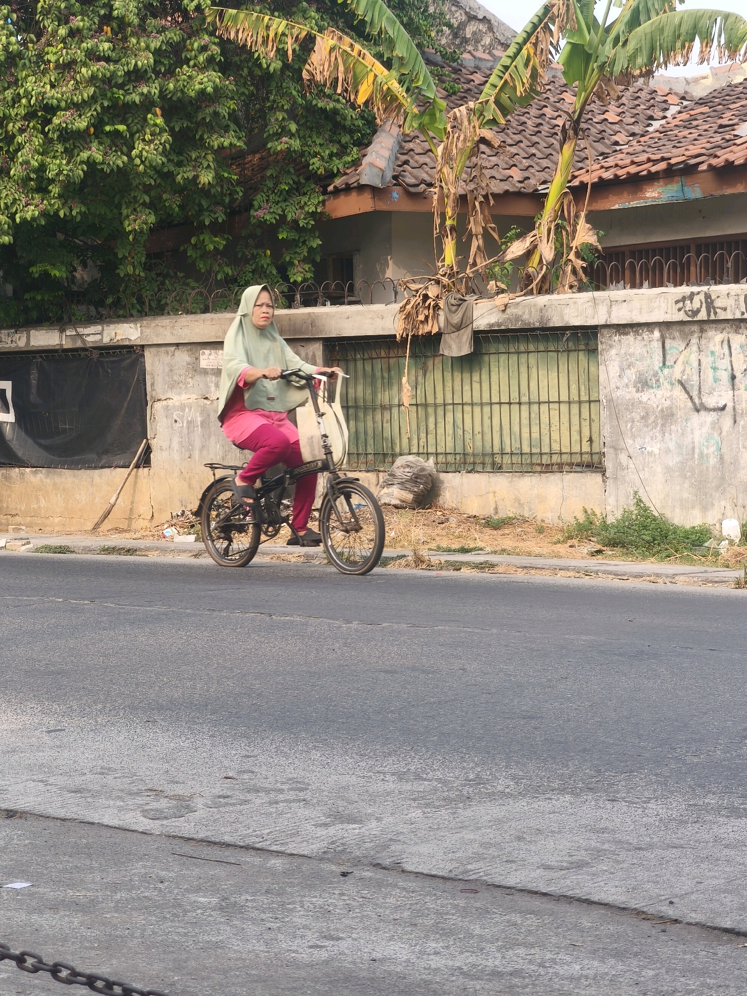 A woman wearing a light green hijab and pink pants is riding a black folding bicycle on a paved road. The bicycle has a white bag attached to the handlebars. The woman appears to be in her 50s or 60s. In the background, there is a concrete wall with graffiti, a green metal gate, and a tiled roof. Banana trees with drying leaves are visible above the wall. The scene is outdoors, and the lighting suggests it is daytime, possibly late afternoon, given the angle of the sun. The location is identified as Bekasi, Indonesia, suggesting an urban or suburban environment.