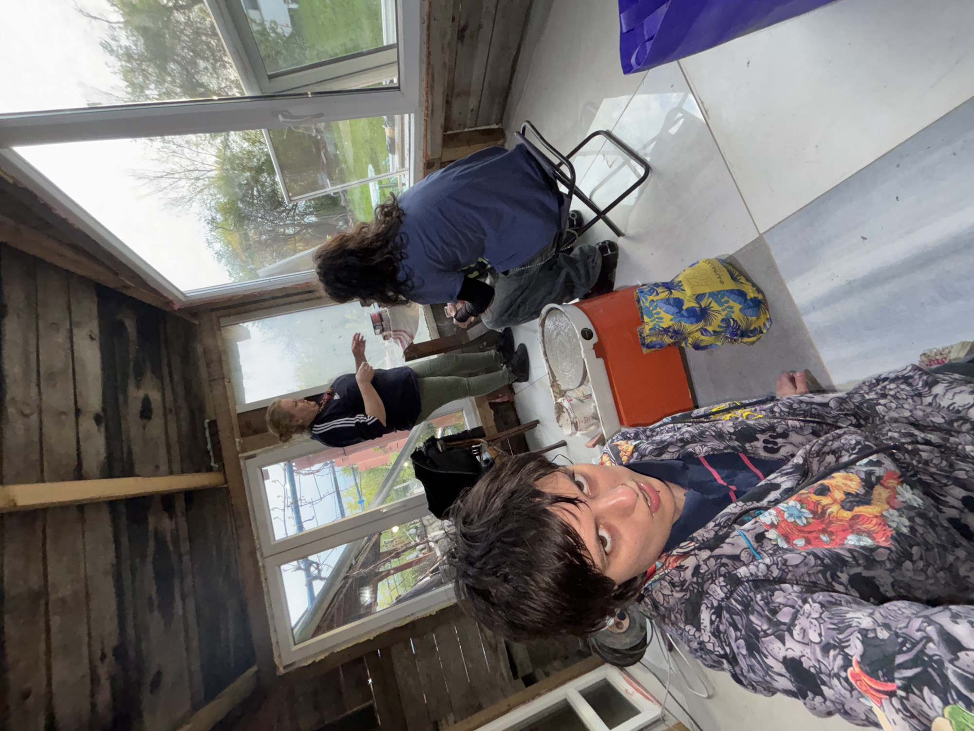 An indoor scene is captured from a low angle, showing three individuals in a room with large windows. The image is oriented sideways, with the floor running vertically on the right side of the frame and a wooden wall on the bottom.
In the foreground, to the right, a person with dark hair, a nose piercing, and a lip piercing is taking a selfie. They wear a dark blue shirt under a black and grey patterned jacket featuring colorful motifs of dragons or similar creatures. Their expression is neutral as they look directly at the camera.
In the middle ground, further back and to the left, another person with long dark hair kneels or crouches on the tiled floor. They wear a blue t-shirt and grey pants and appear to be focused on an activity near a black metal folding stool. An orange bucket and a yellow and blue patterned fabric item, possibly a bag or cloth, are visible nearby. The fabric has partial text, including "V-A.RAKUE" and "CANVAS".
In the background, to the far left, a third person stands, wearing a dark blue shirt with white stripes (possibly athletic wear) and green pants. They have light-colored hair and are gesturing with their hands, seemingly in conversation or instruction.
The room features a light-colored tiled floor and rustic, unfinished wooden walls, suggesting a renovation project or a workshop. Large windows line the left side of the room, offering a view of green trees and bright daylight outside, indicating daytime. A purple bag is also visible on the tiled floor. The overall setting appears functional rather than decorative, potentially in a rural area like Verinsko, Bulgaria, given the external foliage.