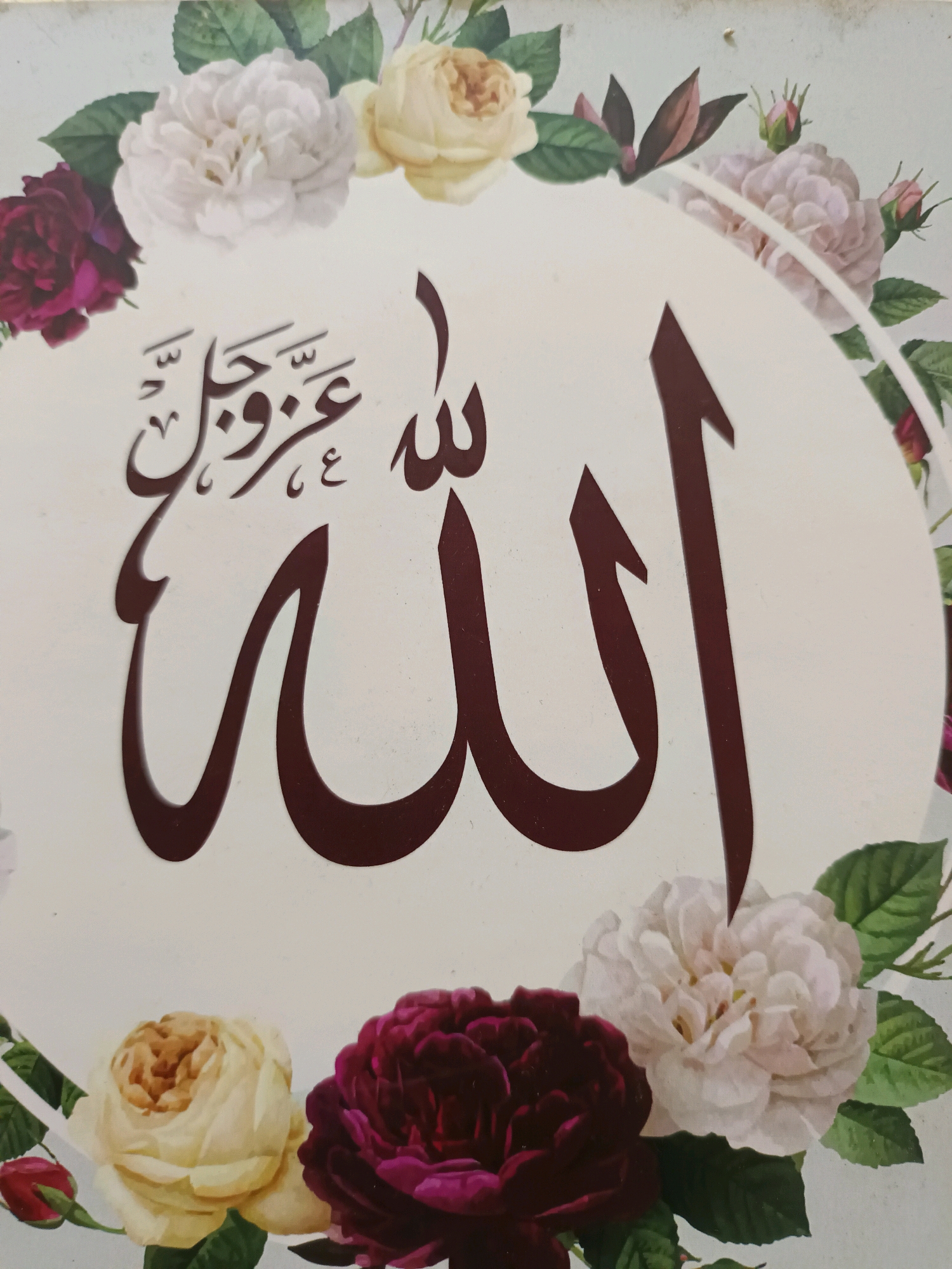 This is a close-up photograph of Islamic calligraphy written in Arabic script against a white background, framed by a floral border of roses in shades of white, cream, and deep red. The calligraphy reads "Allah" in large, bold, dark red letters. Above and to the left of "Allah", there is smaller calligraphy that appears to be "عز وجل" which translates to "the Exalted". The image does not provide any information about its location in Gunung Sekar, Indonesia, nor does it contain any people or interactions. The focus is solely on the decorative calligraphy and floral elements.