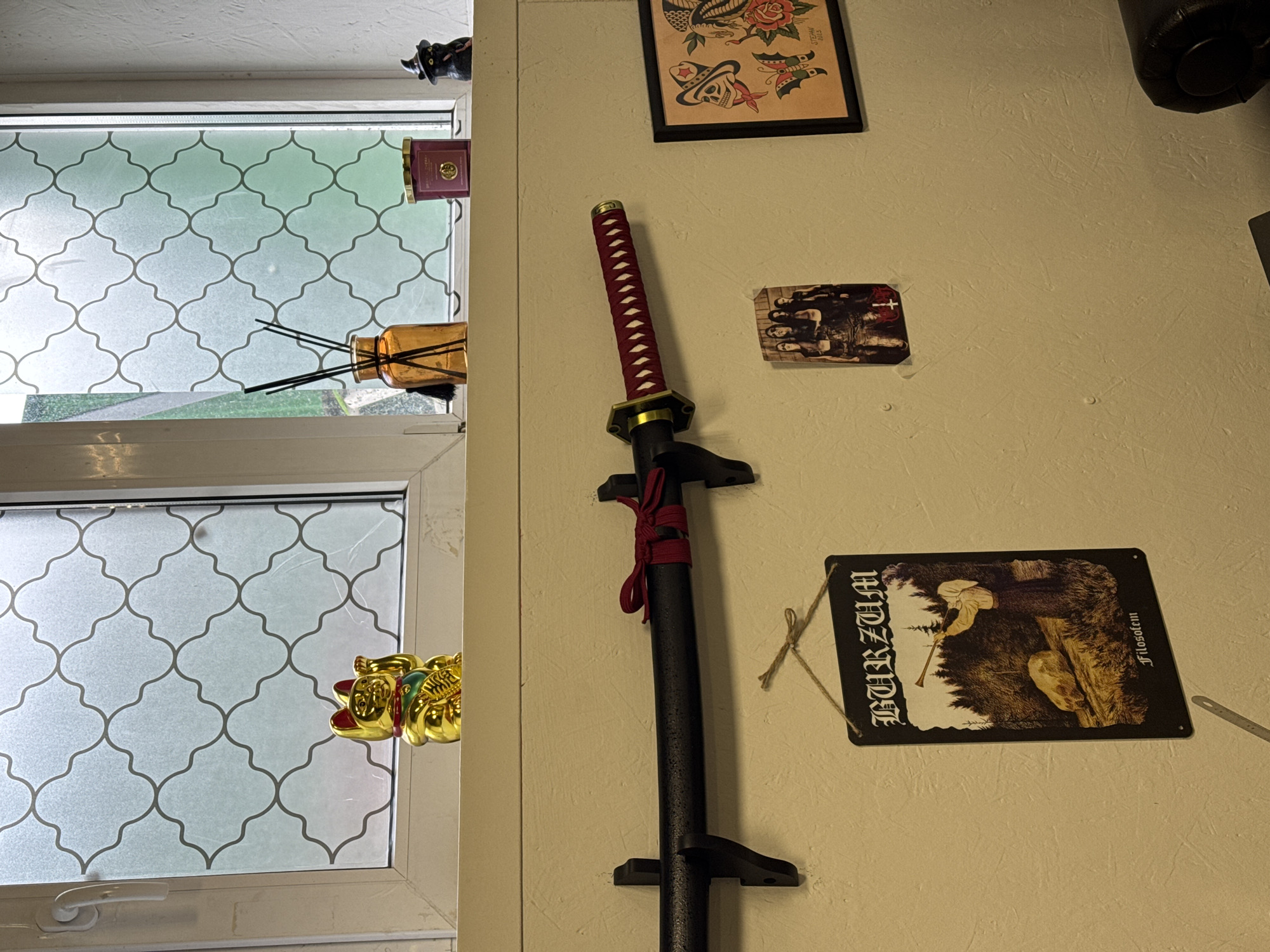 The image shows a room interior in an unknown location. On a light beige wall hangs a katana with a red handle and a black scabbard mounted on a black display stand. Above the katana are various items: a framed tattoo-style print, a small rectangular image affixed to the wall, and a black object near the top right corner of the frame. To the left of the katana is a window with a decorative frosted glass pattern. In the window, one can see an air freshener bottle, a candle, a black cat figurine, and a gold lucky cat statue. The lighting in the room appears to be ambient.