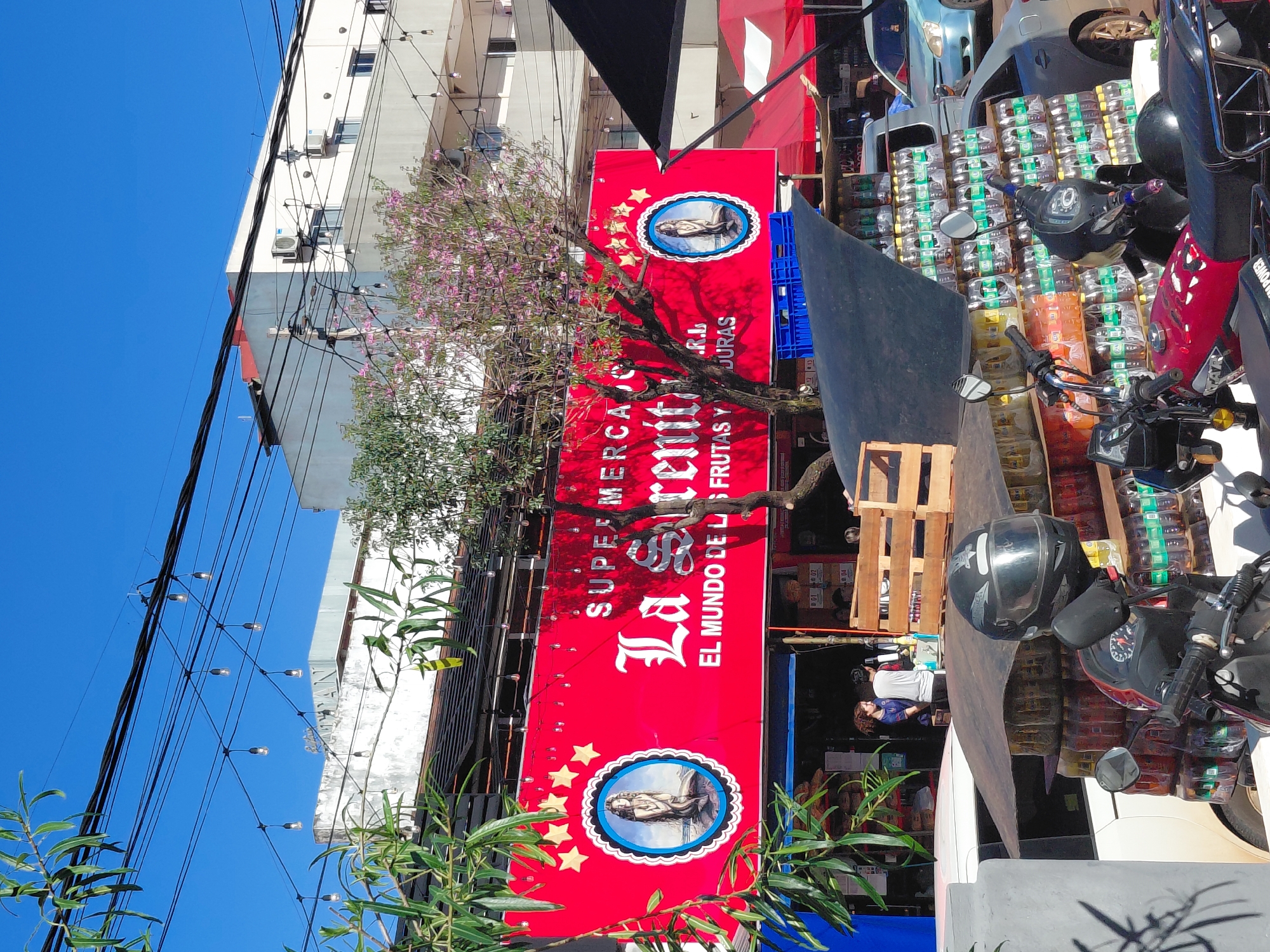 In Encarnacion, Paraguay, a street-level view captures a bustling commercial scene under a clear blue sky. Dominating the frame is a large red banner with white lettering that reads "SUPER MERCADO La Serenitiima" and below it, "EL MUNDO DE LAS FRUTAS Y VERDURAS". Two circular emblems, one on the left and one on the right of the banner, feature a depiction of a woman. The banner is mounted on the facade of a building, with the upper floors of other buildings visible behind it.
In front of the building, various items are displayed for sale. Stacks of plastic bottles filled with liquid, possibly beverages, are arranged on wooden shelves and pallets. A black tarp is partially covering some of these goods. A motorcycle with a black helmet on its handlebars is parked in the foreground, partially obscuring the view of the merchandise. A white vehicle is parked behind the motorcycle.
In the background, people are seen interacting near the entrance of the supermarket, with one individual appearing to be a cashier. String lights are strung across the street above the banner, and overhead wires crisscross the sky. Further in the background, a light blue car is visible parked on the street. The overall impression is of a vibrant outdoor market or retail area in a South American city.