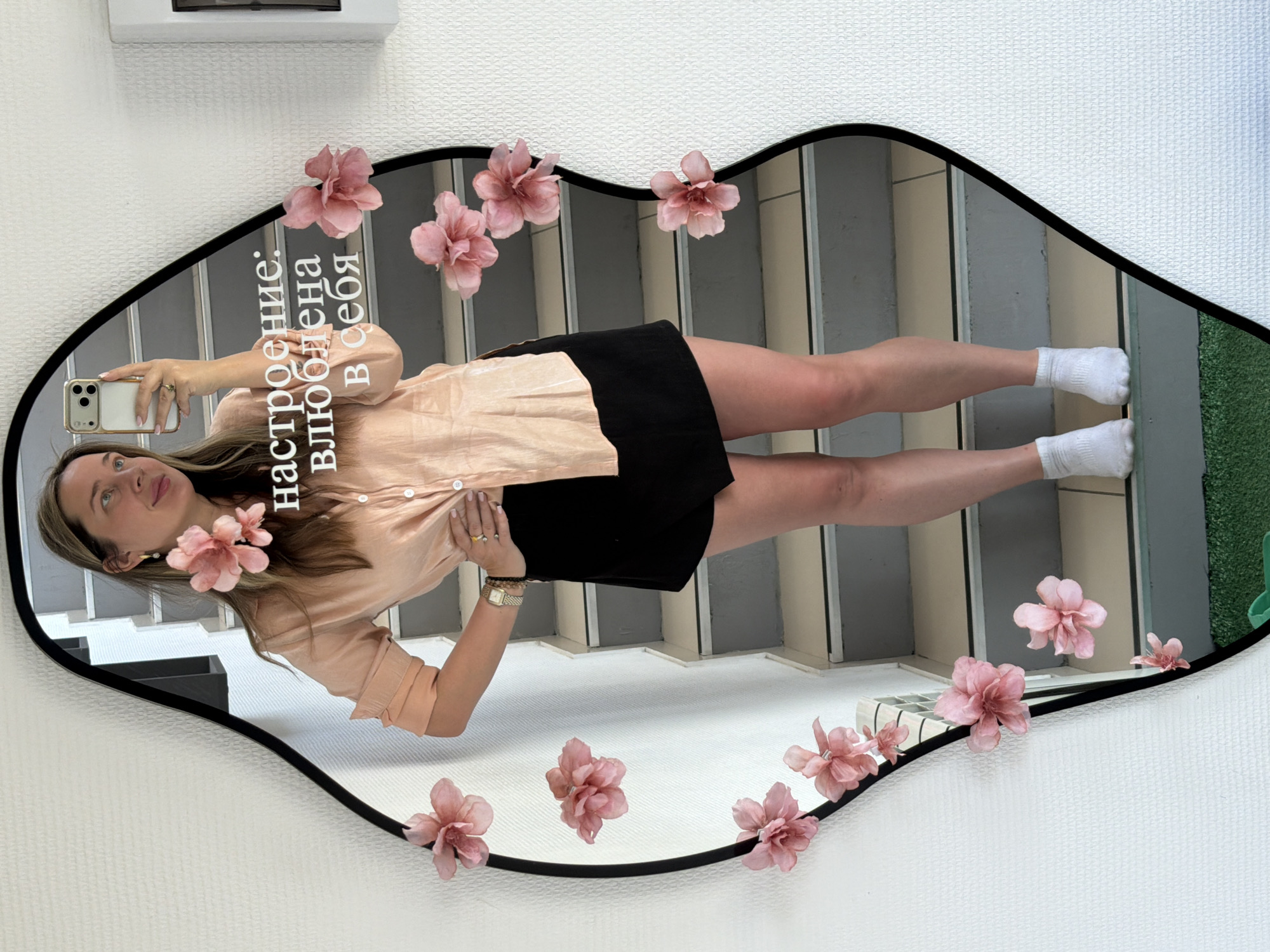 A person is reflected in an ornate mirror decorated with pink flowers. The person is holding a smartphone, presumably taking a selfie. The mirror is hanging on a textured white wall. The reflection shows a staircase and a strip of artificial grass. The text on the mirror is in Russian and says "настроение: настроена влюблена в себя", which translates to "mood: настроена in love with herself." There is a white box above the mirror. The location is unknown, including whether the image was taken in Chaikovsky, Russia.