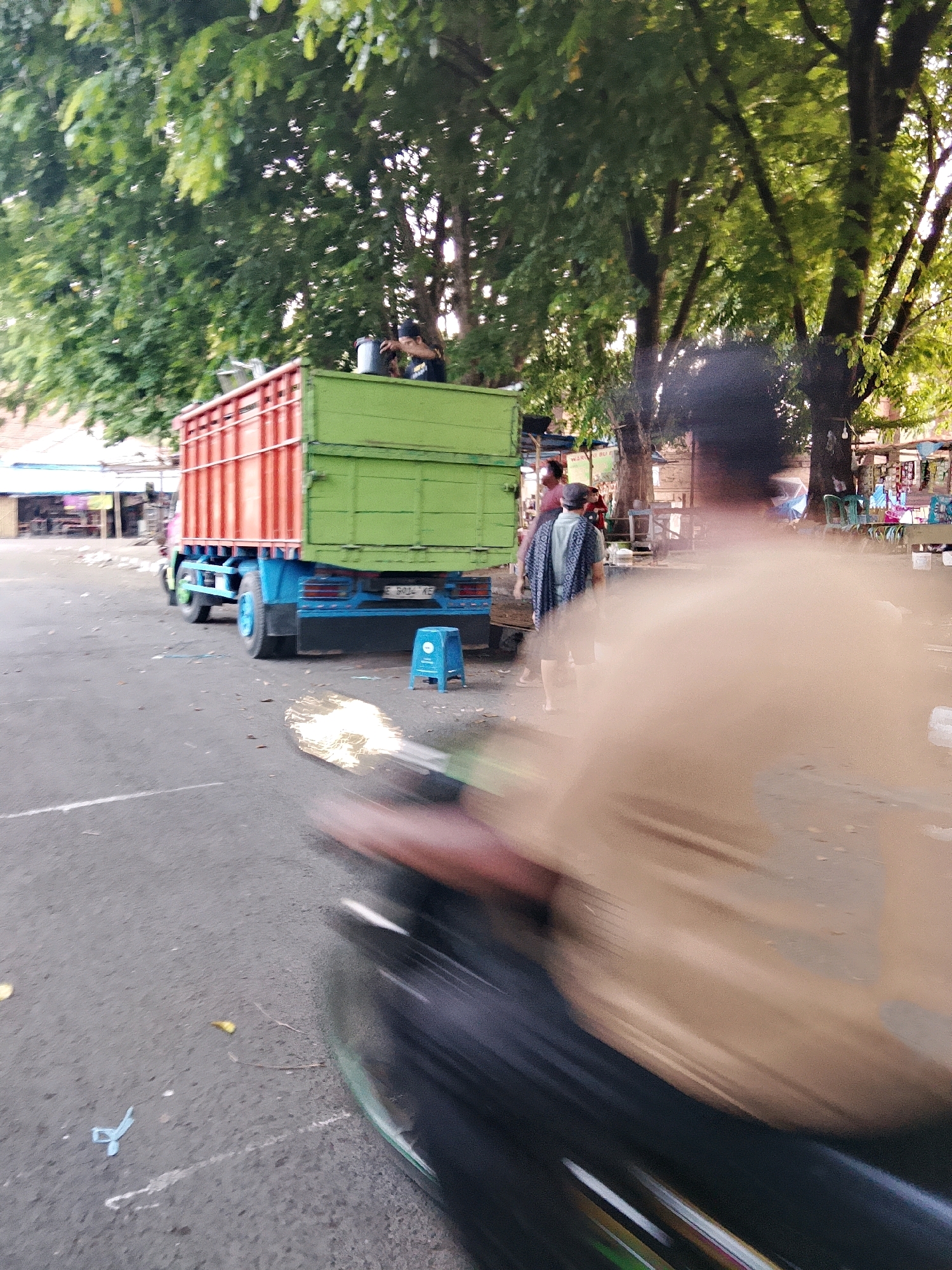 The image captures a scene outdoors in Cirebon, Indonesia. A blue and green truck with an orange-railed cargo bed is parked on an asphalt surface. To the right of the truck, a small blue stool is visible. In the background, there are trees with lush green foliage, and structures that appear to be market stalls or shops with awnings. Several individuals are present. One person, wearing a patterned shirt and shorts, is standing near the truck. Another person is seated on top of the truck, holding a bucket. The foreground is dominated by a blurred, motion-captured view of a person on a motorcycle, with their arm and torso visible, suggesting movement. The lighting indicates it is daytime, likely morning or late afternoon given the soft sunlight filtering through the trees.
