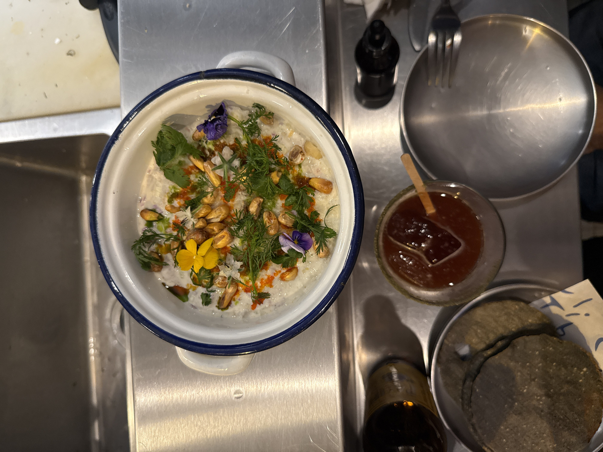 The image shows a close-up, overhead view of a meal preparation on a stainless steel surface, potentially a kitchen counter or sink. The main subject is a white bowl with a dark blue rim, containing a creamy-looking dish adorned with edible flowers, herbs, and what appear to be toasted corn kernels. Next to the bowl is a small bottle of what appears to be hot sauce or other condiment, a plate with utensils, and a small glass bowl containing a reddish-brown sauce with a wooden spoon or stirring stick inside. Also visible is a small stack of what appear to be tortillas in a dish, and another small bottle filled with a dark liquid.
While I can't definitively pinpoint the location as Mexico City without additional information, the ingredients and presentation of the food, particularly the use of corn and the presence of what could be a salsa-like sauce, suggest Mexican cuisine. Mexico City is known for its diverse food scene, so a dish like this would not be out of place.