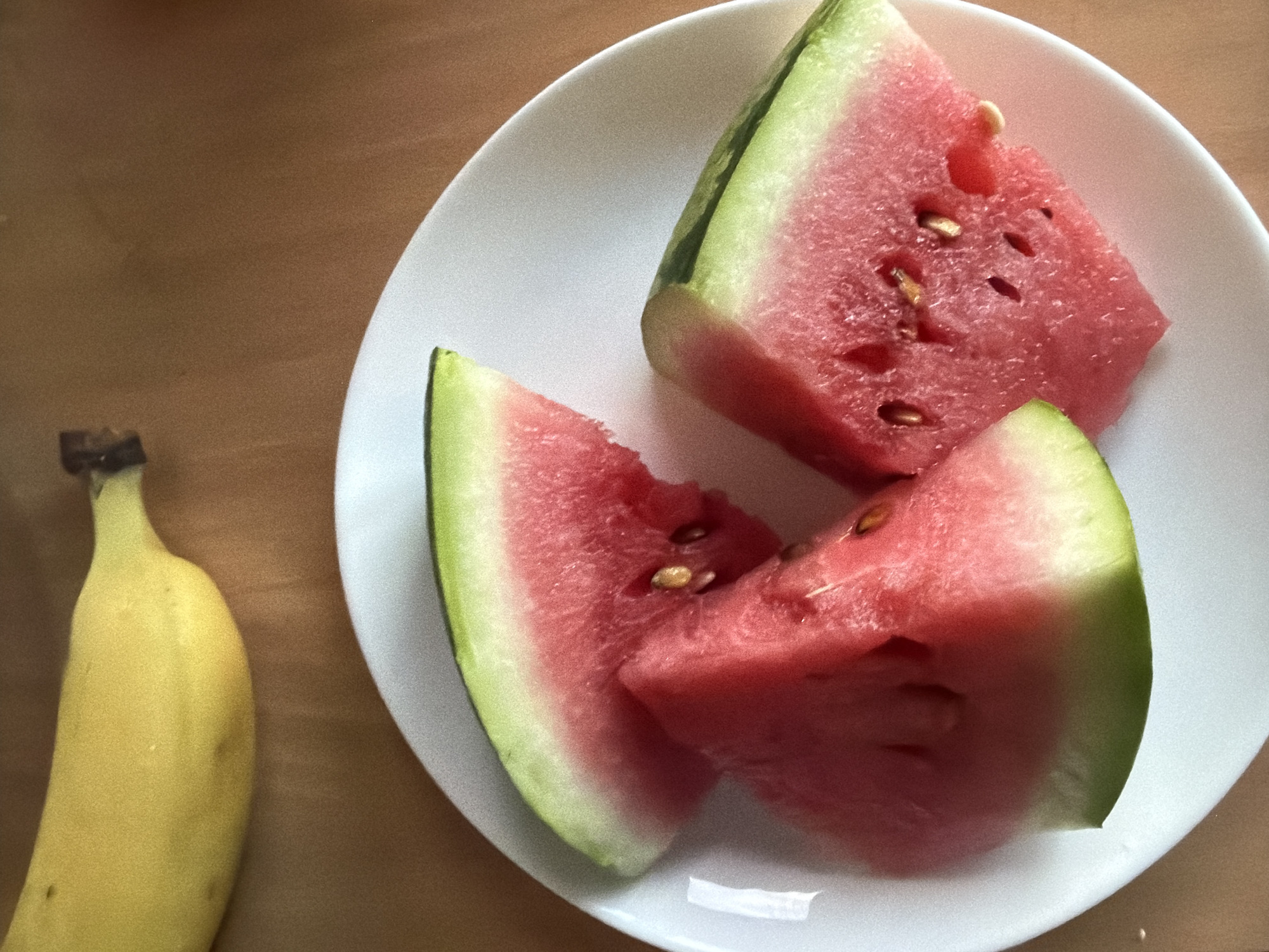In an overhead shot, a bright yellow banana is positioned in the upper left corner of the frame, resting on a brown surface. To the right of the banana, a white plate holds three triangular slices of watermelon. The watermelon pieces are a vibrant pink with visible seeds. The overall scene suggests a simple meal or snack.