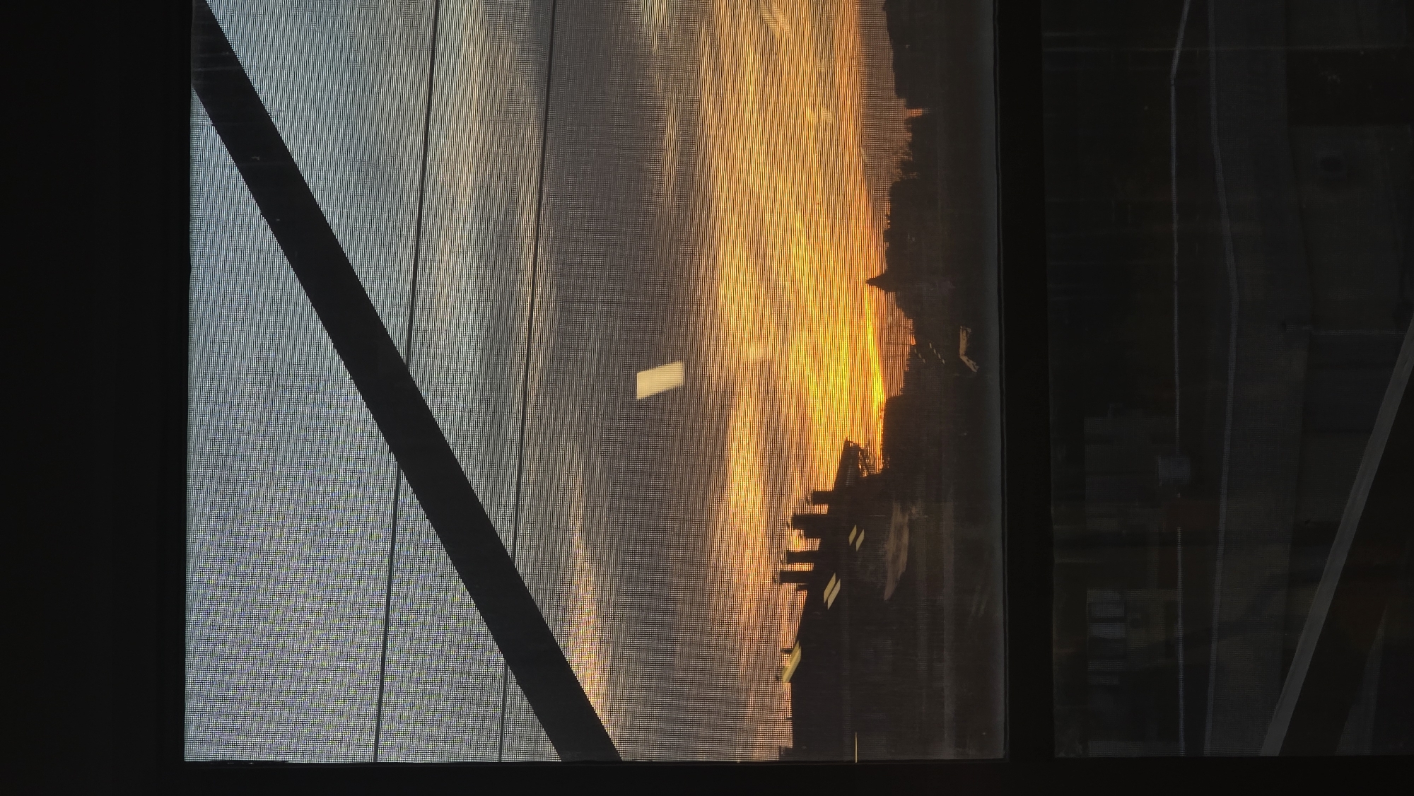 The image, taken in Sofia, Bulgaria, captures a vivid sunset or sunrise viewed through a window screen. The scene is dominated by a dramatic sky, split diagonally by a dark, structural beam or window frame. To the left, the sky is a pale, cool blue, gradually transitioning to a brilliant palette of orange and yellow hues towards the right horizon, indicative of the sun's position.
Against the glowing sky, the dark silhouettes of a city or urban landscape are visible along the horizon line. These include various building structures with distinct rooflines and projections. The entire outdoor view is filtered through a fine, grid-like pattern, characteristic of a window screen or mesh, which is consistently visible across the illuminated sky.
A prominent, dark, diagonal bar extends from the upper left corner downwards and rightwards across the window, obscuring a portion of the sky. In the central right area of the bright orange sky, a distinct, rectangular, glowing white shape is visible, possibly a reflection or an illuminated distant object. The far right side of the image is very dark, possibly showing an interior space or a heavily shadowed exterior, with faint vertical lines suggesting further structural elements.
The time of day is clearly either early morning or late evening, given the warm, intense colors of the sky. The weather appears clear. No visible text is present in the image. The composition suggests a moment of tranquil observation from indoors.