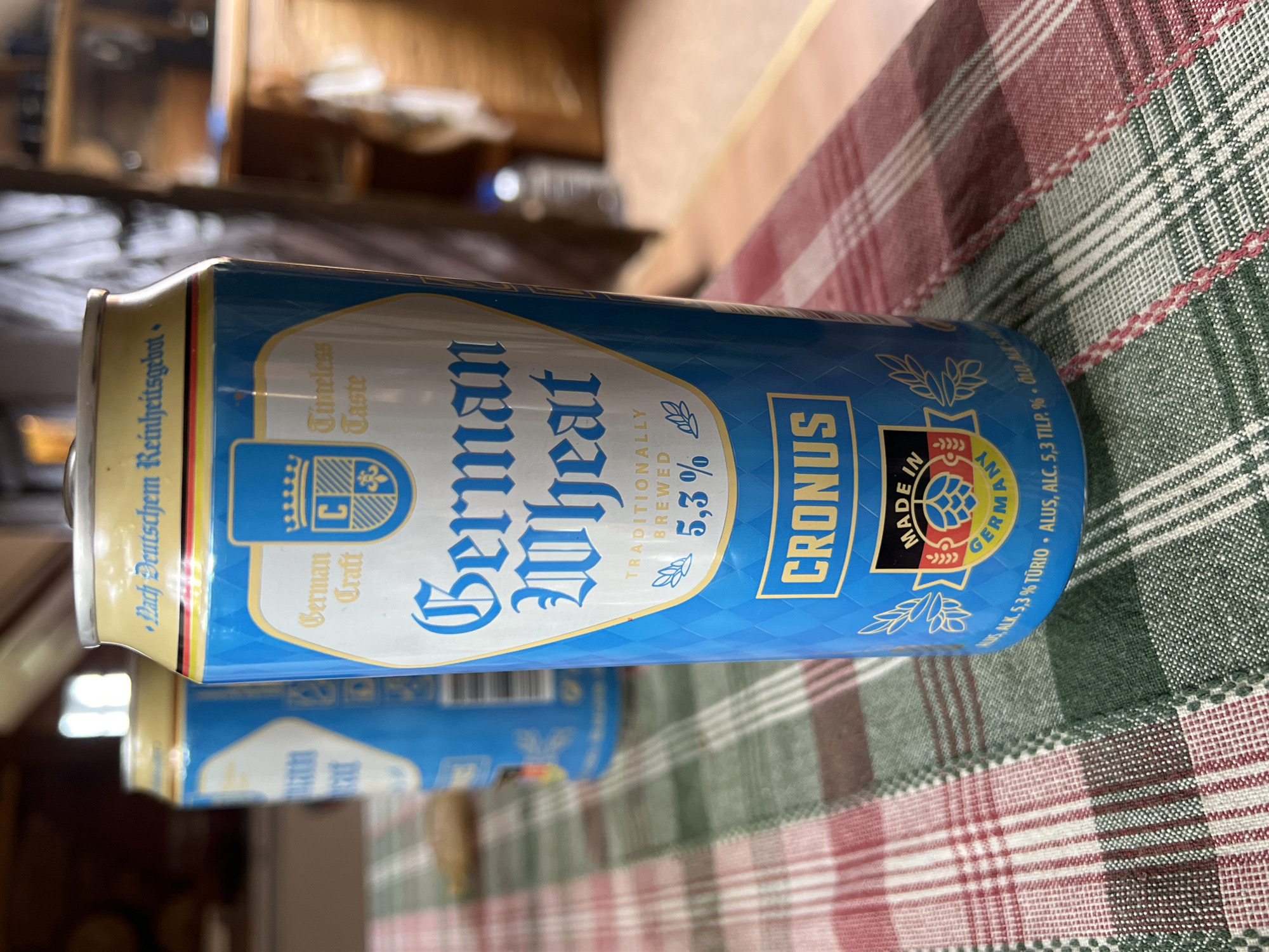 A close-up, eye-level, medium shot shows two blue beer cans with yellow and red accents on a red, white, and green checkered tablecloth. The can in the foreground is a Cronus German Wheat beer can, displaying text such as "German Craft", "Timeless Taste", "TRADITIONALLY BREWED", "5,3%", and "MADE IN GERMANY". The can in the background is also a blue beer can with yellow and red accents. The image is focused on the beer cans, with the background out of focus, showing glimpses of wooden furniture and a wall. The scene is set indoors.