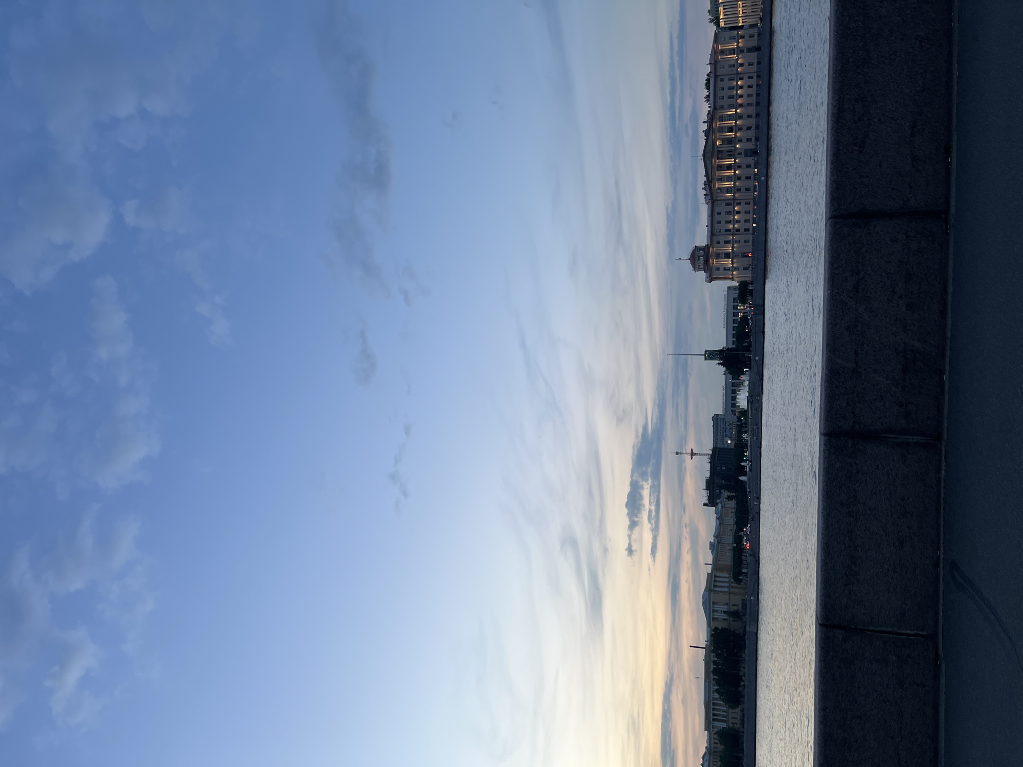 A wide shot captures a city skyline across a body of water at dusk. The sky is a blend of blues and soft oranges, with scattered clouds. On the far bank, buildings of various architectural styles line the waterfront. One prominent building, illuminated by lights, features classical columns and a domed roof. To the left, a tall, spire-like structure is visible, along with other less distinct buildings. Trees are scattered between the structures. The water in the foreground reflects the muted colors of the sky and the lights of the city. A thick, stone embankment runs along the bottom of the frame, with a dark asphalt surface in front of it. The scene suggests a tranquil evening in Saint Petersburg, Russia, likely along one of its many canals or the Neva River. No individuals are clearly discernible.