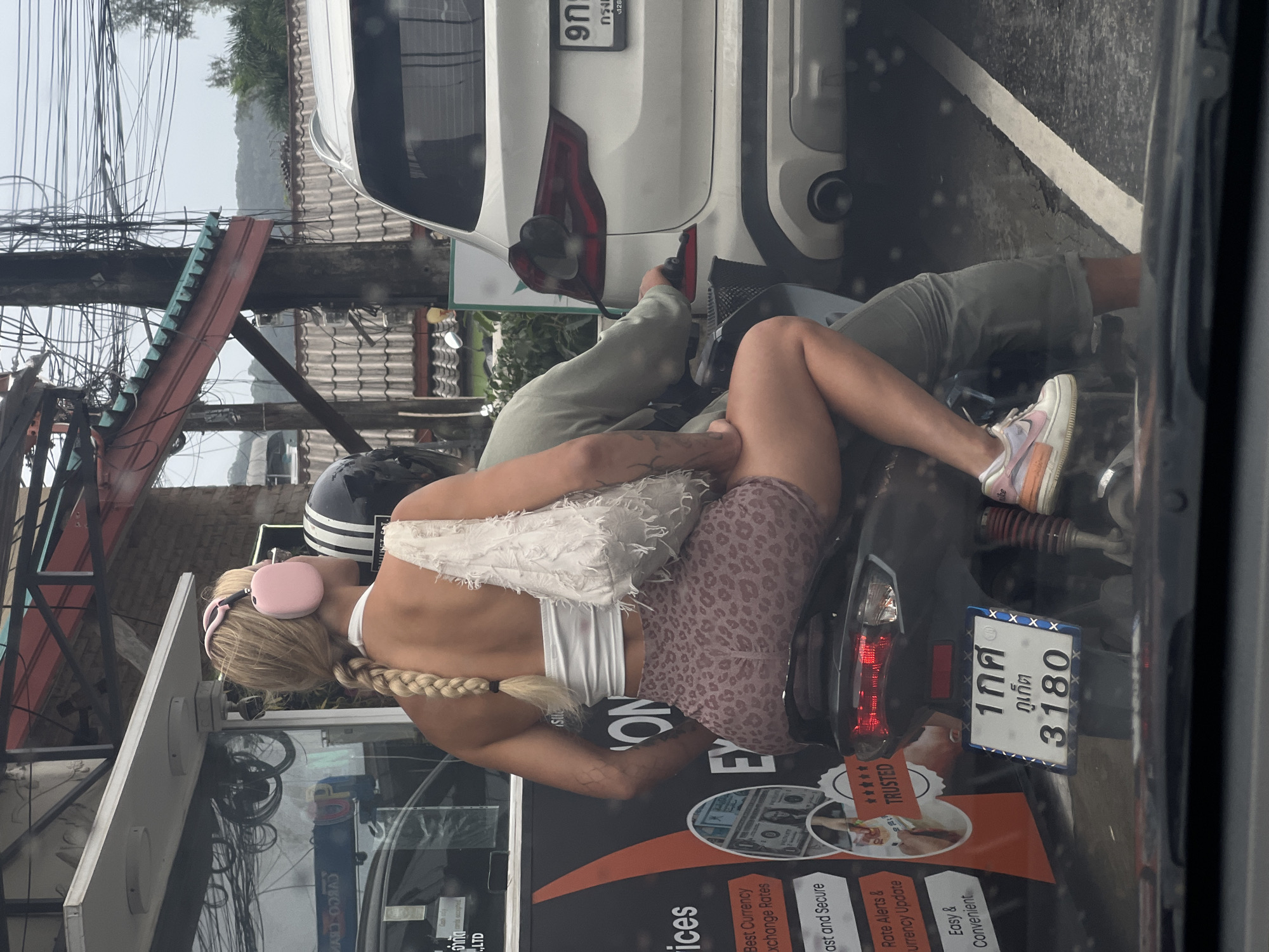 Here is a detailed description of the image:
**Scene:** A photograph, likely taken from inside a vehicle, captures a scene on a busy street in what appears to be a Southeast Asian urban area. The image is focused on a scooter carrying two individuals.
**People:**
* A blonde woman with a long braid is riding on the back of the scooter. She is wearing a light-colored top, a patterned skirt, and pink headphones. She has tattoos on her left arm.
* The scooter driver is wearing light-colored pants and sneakers.
**Objects:**
* A black and gray scooter with a Thai license plate visible. The plate reads "1กศ ภูเก็ต 3180". The scooter has advertising stickers on it.
* A white car is parked close to the scooter.
* A motorcycle helmet is visible.
* There is a street sign on the left. The sign has a logo visible and some information written in English.
* Overhead, there are many tangled electrical wires.
**Environment:**
* The street is lined with buildings and appears to be relatively narrow.
* The image suggests a densely populated area.
**Actions and Interactions:**
* The two people are riding on the scooter, likely in traffic.
* The woman on the back of the scooter seems relaxed.
**Location:**
* Based on the Thai license plate, the location is likely in Phuket, Thailand.