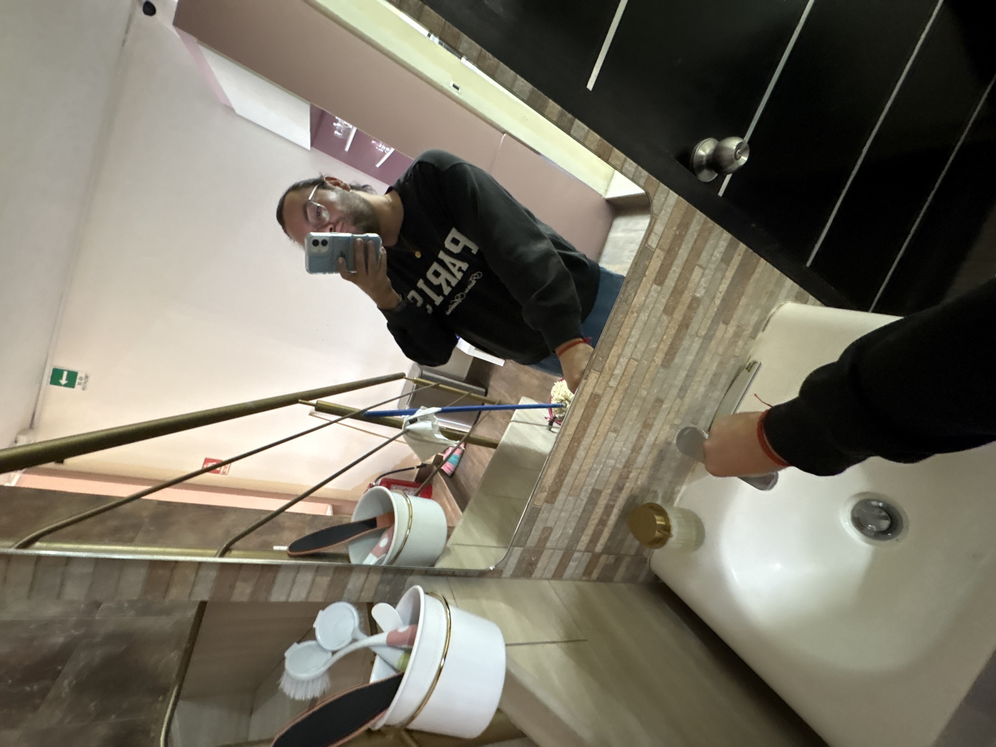 A mirrored selfie taken in a bathroom. A man with glasses and a black sweater is taking the photo with his phone. The sink is white and the wall behind it is tiled. The reflection shows the upper half of stairs with a bronze railing, a basket of grooming supplies, and a green exit sign. The location is likely in San Bernardino Tlaxcalancingo, Mexico.