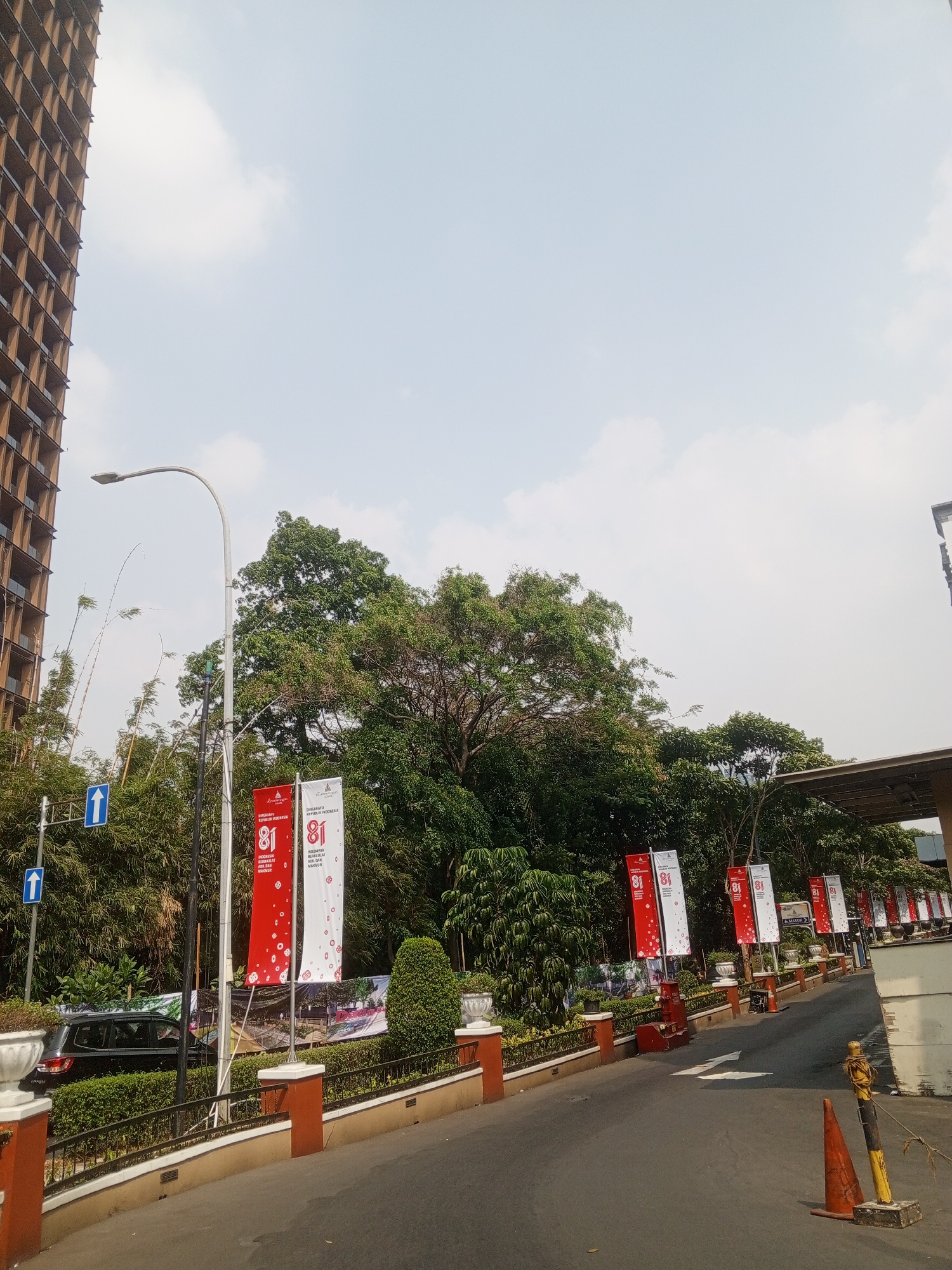 This image shows a street scene in South Jakarta, Indonesia, on a clear day with scattered clouds. On the left, a tall, multi-story building with a visible grid pattern of balconies dominates the background. A street light with a curved arm is positioned in the mid-ground. In front of the building and extending towards the center of the image are dense green trees.
Along the right side of the road, several vertical banners are displayed. These banners are predominantly red and white, featuring the number "81" prominently. Some also have text in Indonesian. The banners are hung from poles and spaced along a sidewalk area.
The road itself is paved and appears to be a thoroughfare. There are lane markings, including a white arrow indicating direction. To the right of the road, there is a building with a covered walkway or entrance. A few orange traffic cones are visible, one in the foreground and another further down the road. In the mid-ground, a dark-colored car is parked near the sidewalk. The environment appears to be a developed urban area with landscaping elements like hedges and decorative planters.