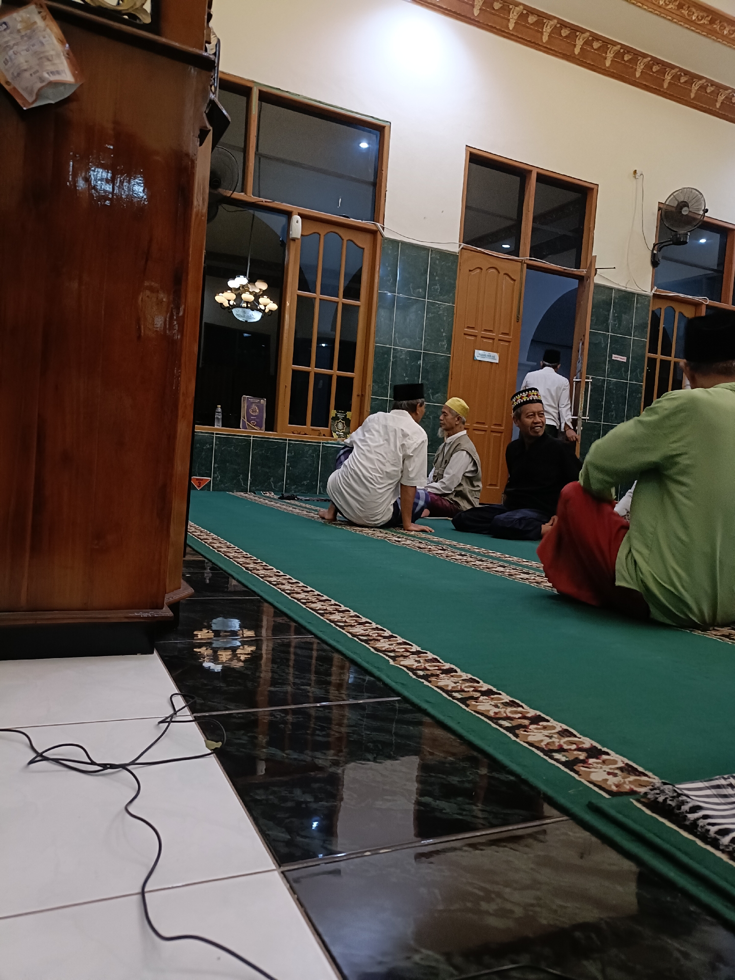The image depicts the interior of a mosque in Kediri City, Indonesia, likely during prayer or a gathering. Several men are seen sitting on a green carpet, some wearing traditional Islamic attire. One man in a black shirt and a patterned cap is smiling and appears to be engaged in conversation with others. Another man in a green shirt and red sarong is seated facing away from the camera. The mosque features wooden doors, windows with dark panes, and tiled flooring. A large wooden cabinet is visible on the left side of the frame, and a fan is mounted on the wall. The lighting suggests an indoor setting, possibly late afternoon or evening.