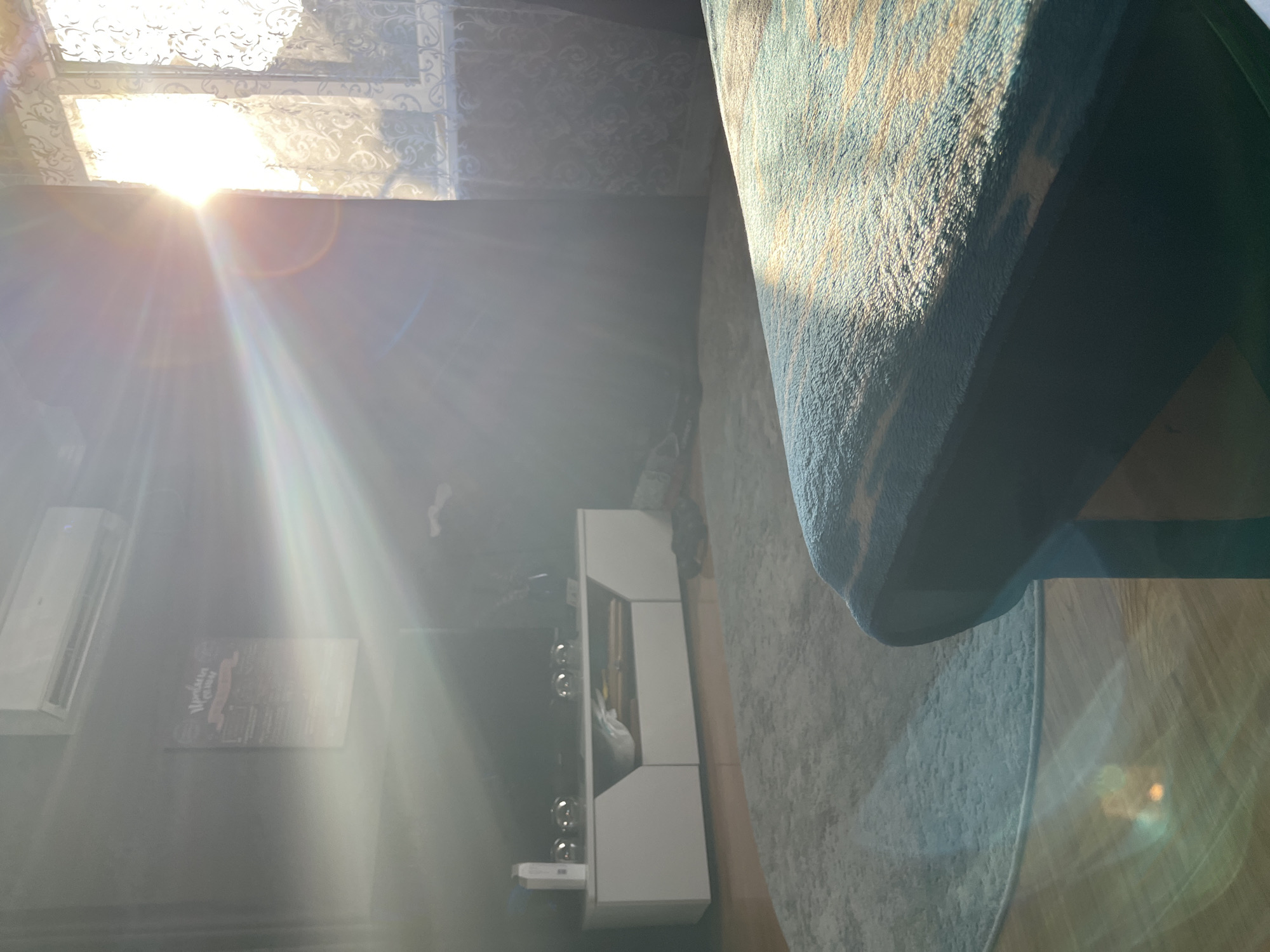 The image depicts an indoor scene, likely a living room, with strong natural light streaming in from a window on the right, creating lens flares and a bright, hazy atmosphere. A white media console with shelving and cabinets is visible on the left, with some decorative items and a white bottle on top. A circular rug covers a portion of the wooden floor in front of the console. In the foreground, the edge of a dark-colored piece of furniture, possibly a sofa or ottoman, covered with a teal and beige patterned blanket, occupies the lower right portion of the frame. The wall on the left is dark-colored and features an air conditioning unit at the top and a framed poster with text in Cyrillic script. The scene is devoid of any individuals or discernible actions. The location is identified as Saratov, Russia.