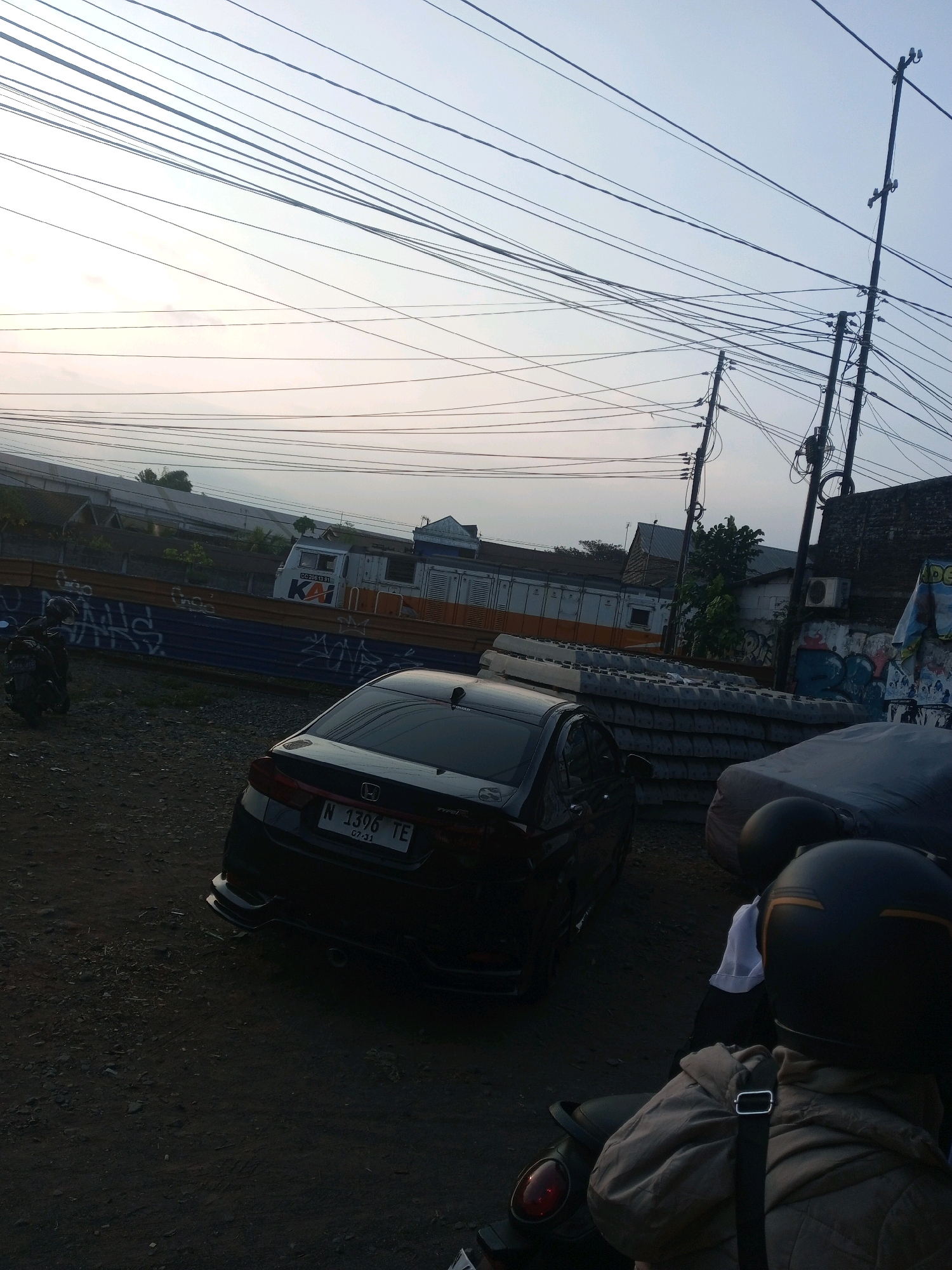 The image depicts a street scene, likely in Malang, Indonesia, given the license plate of the car, which reads "N 1396 TE". The time of day appears to be late afternoon or early evening, with the sky exhibiting a soft, diffused light. Overhead, a complex network of electrical wires crisscrosses the frame, supported by utility poles.
In the foreground, a black car is parked, its rear visible to the viewer. A motorcycle is parked to the left of the car, with its front wheel partially obscured. Further in the background, a train with "KA" markings is visible, suggesting proximity to a railway line or station. The environment appears to be a mix of urban and industrial, with buildings, a fence with graffiti, and stacks of what appear to be concrete or construction materials. There are no clearly visible people engaged in any specific actions, though a helmet and part of a jacket are visible on a motorcycle in the lower right quadrant, indicating the presence of at least one individual, likely on the motorcycle. The ground is unpaved, covered with gravel and dirt.