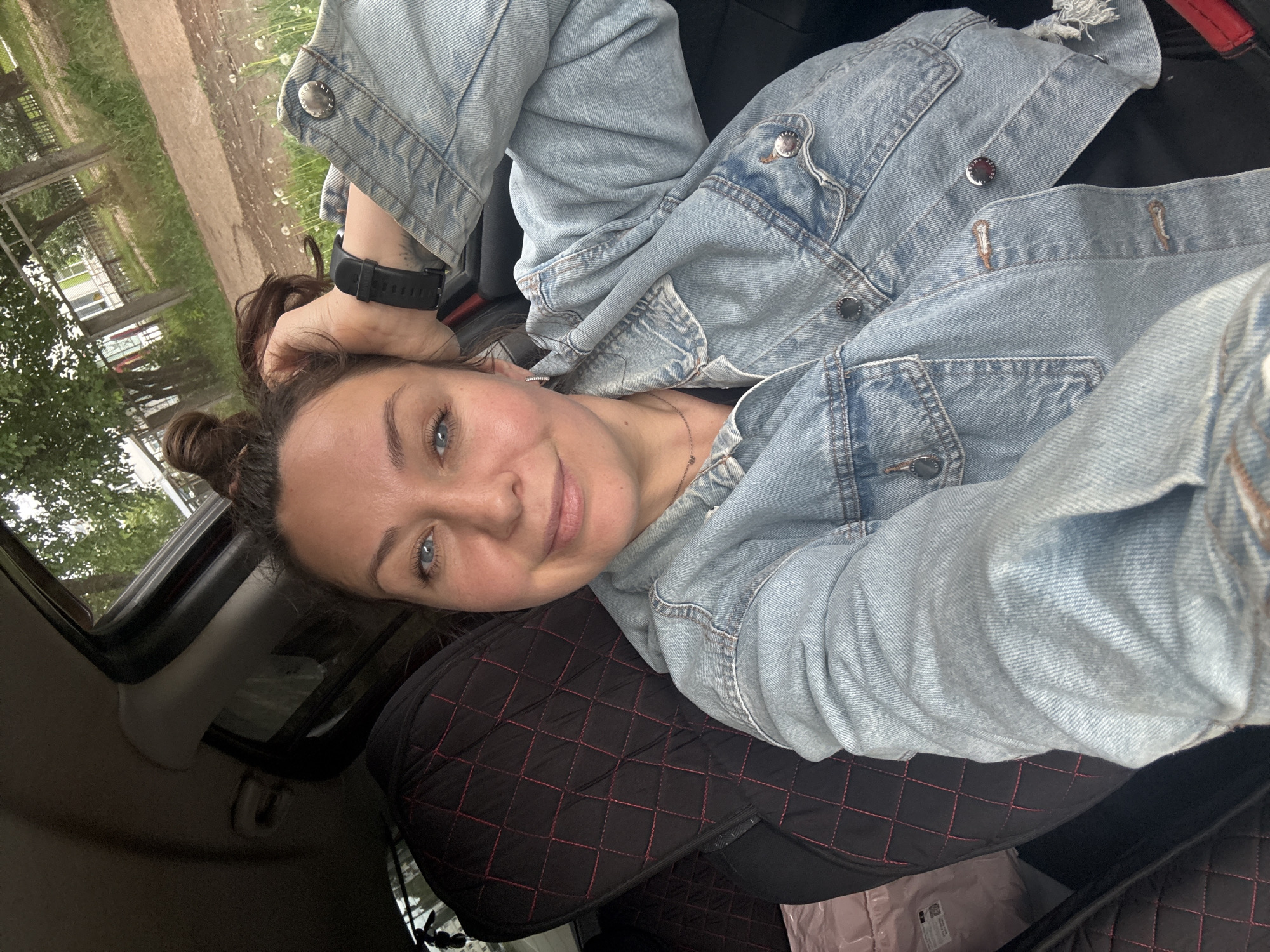 An adult female, estimated to be in her 30s-40s, is seated in the driver's seat of a vehicle in Chaikovsky, Russia, taking a selfie. She looks directly at the camera with a soft smile. Her dark brown hair is styled in a messy top knot, and she has clear blue eyes, full eyebrows, and fair skin. She wears a light-wash denim jacket with multiple metallic buttons and breast pockets over a dark top, a thin silver necklace, and a small silver stud earring in her visible left ear. A black digital smartwatch is on her left wrist, and a tattoo is partially visible on her inner forearm. Her left hand is casually resting on her head, touching her hair.
The vehicle's interior features black, diamond-quilted seat covers with red stitching on both the driver's and the visible passenger's seat. A light pink or beige object, possibly a bag, rests on the passenger seat. The dark headliner and window frame are visible above her.
Through the vehicle's windows, an overcast sky and a semi-rural outdoor environment are visible. The background shows mature green trees, lush grass interspersed with white dandelions, a rustic wooden fence with wire, and an unpaved dirt road or path. In the distance, a light-colored building with a green roof is partially obscured by the foliage.