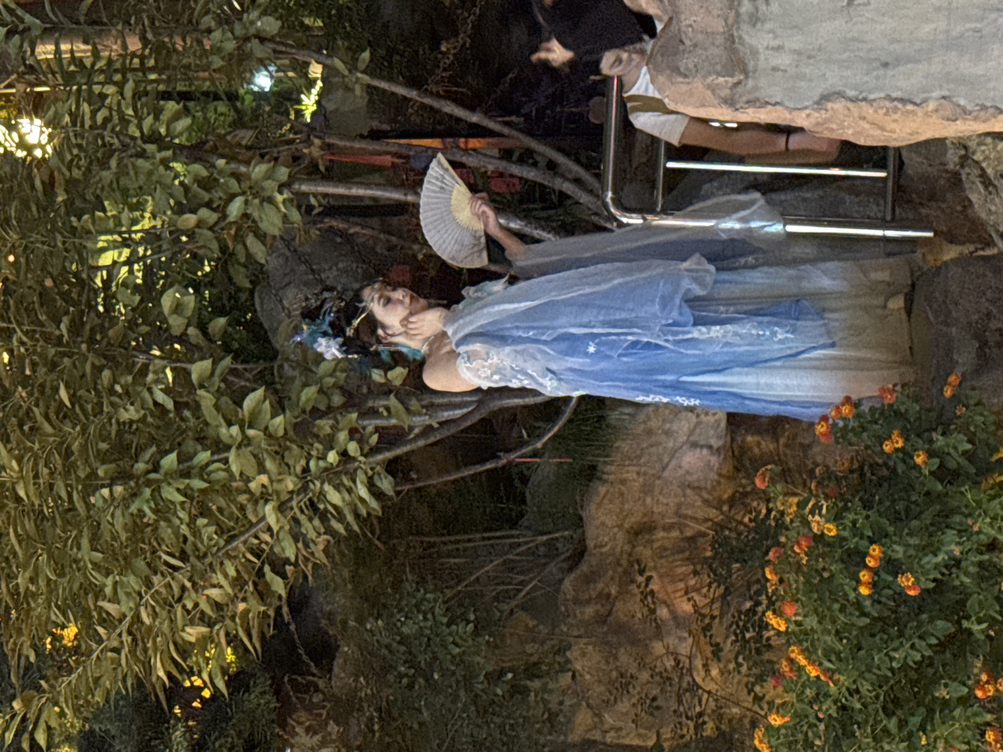 In Yuzhong District, China, a woman dressed in traditional blue Chinese attire stands near a railing, holding a fan. She is adorned with an ornate headpiece and appears to be posing. Behind her, trees and rocks form a naturalistic backdrop, with some people visible in the background. The scene appears to be outdoors, possibly in a park or cultural garden, with soft lighting suggesting it might be evening.