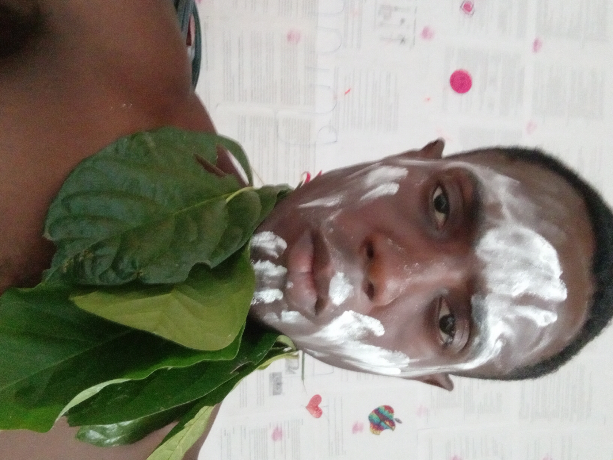 A close-up, rotated image shows a dark-skinned man with white cream or paste smeared across his forehead, cheekbones, and nose. He has short, dark hair. A cluster of large, green leaves is positioned around his neck and shoulder. The background consists of a patterned surface with visible text and scattered pink and multi-colored heart and apple-shaped designs. The location is Akwete, Nigeria.