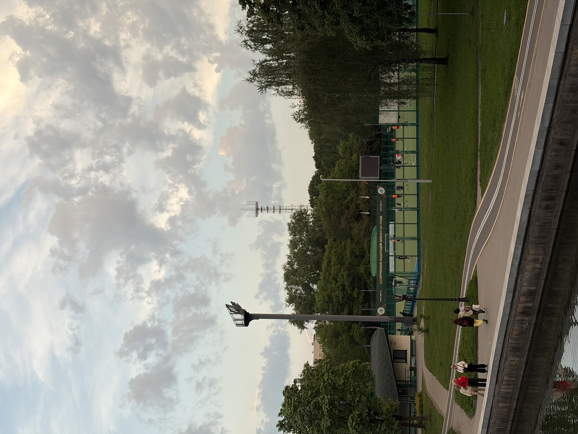 The image depicts a partially overcast sky above a park in Minsk, Belarus. A radio tower is visible in the distance. In the midground, there is a sports complex with fenced-in field hockey pitches. Floodlights stand tall near a small building. A sidewalk runs along the water's edge, where groups of people are walking. The water reflects the sky and the park.