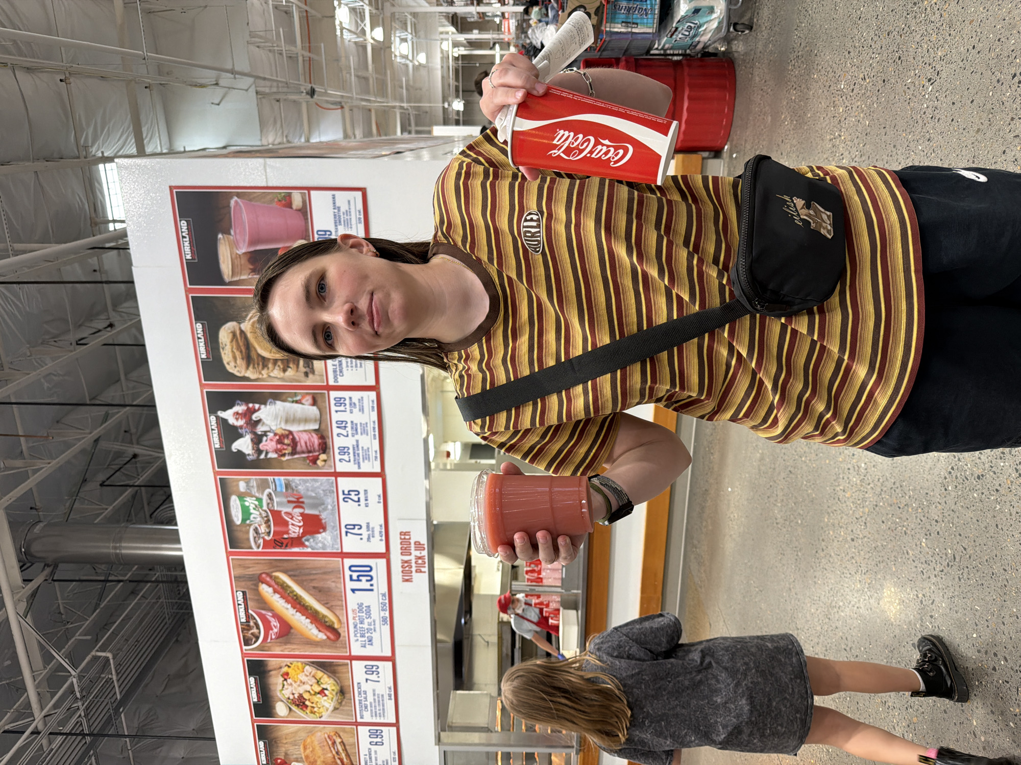 A woman in a striped shirt and fanny pack stands in a Costco food court in Beaverton, Oregon. She holds a red cup of pink smoothie in her left hand and a can of Coca-Cola in her right. In the background, a young girl walks away from the camera, and a Costco employee is visible behind the counter. The food court is brightly lit, with a menu board displaying various food items.