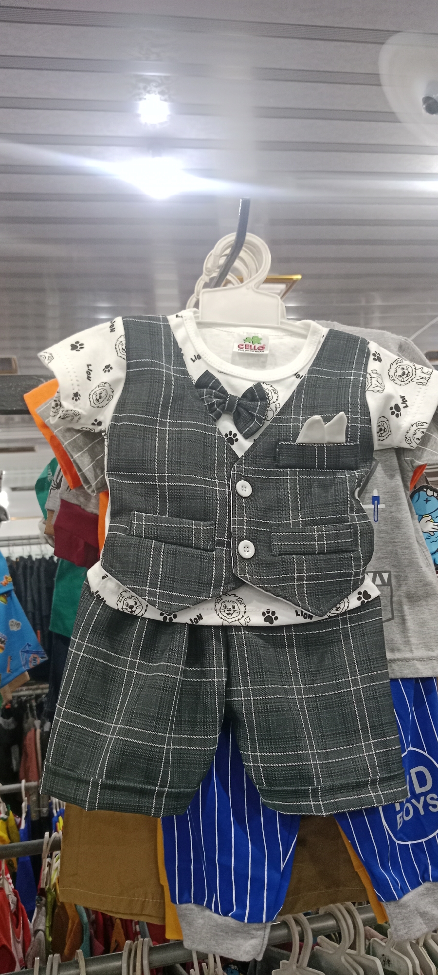A close-up, eye-level shot shows a baby's grey plaid suit with a white t-shirt featuring lion and paw prints. The suit has a bow tie and a pocket square. It hangs on a white plastic hanger on a rack in a clothing store, likely located in Rengel, Indonesia, given the context of previous images. The background shows other children's clothing items also on hangers. The lighting is bright, creating some glare on the ceiling.