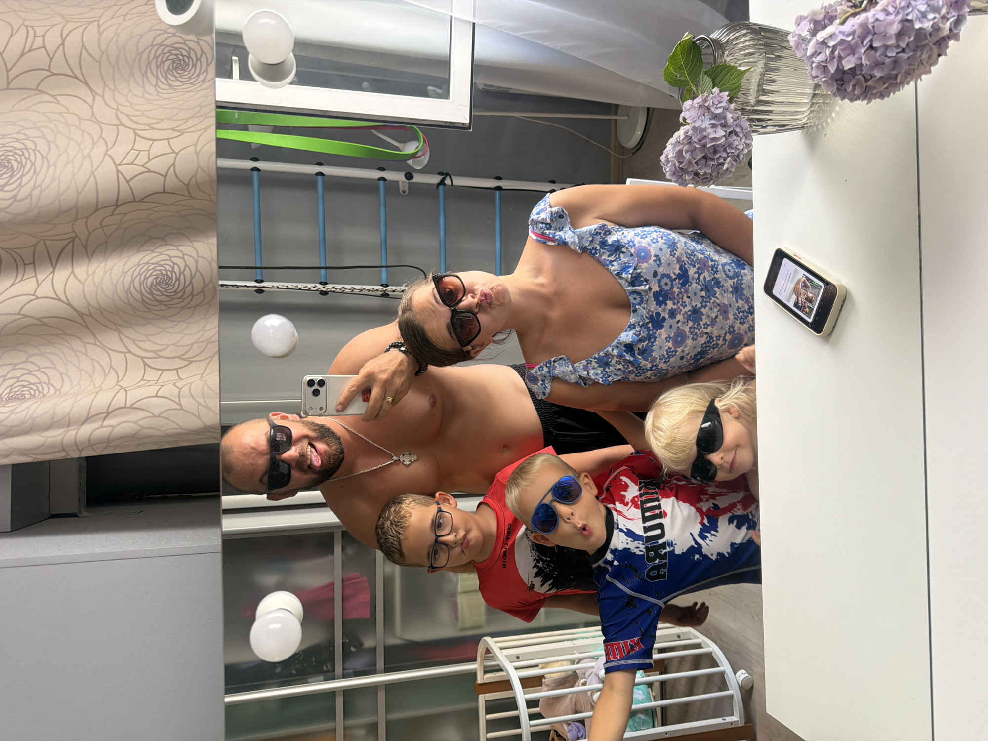 A family of five, two adults and three children, take a mirror selfie in what appears to be a home gymnasium. The father is shirtless and wearing black shorts and sunglasses, sticking his tongue out at the camera. The mother is wearing a blue floral dress and sunglasses, puckering her lips. Two older boys and a younger girl are also in the frame, all wearing sunglasses. The background features a gymnastics ladder, a crib, and decorative curtains. The image is taken at an angle, with the bottom portion of the frame showing a white table with a vase of hydrangeas and a smartphone. The overall impression is a candid, playful family moment. The location is identified as Sochi, Russia.