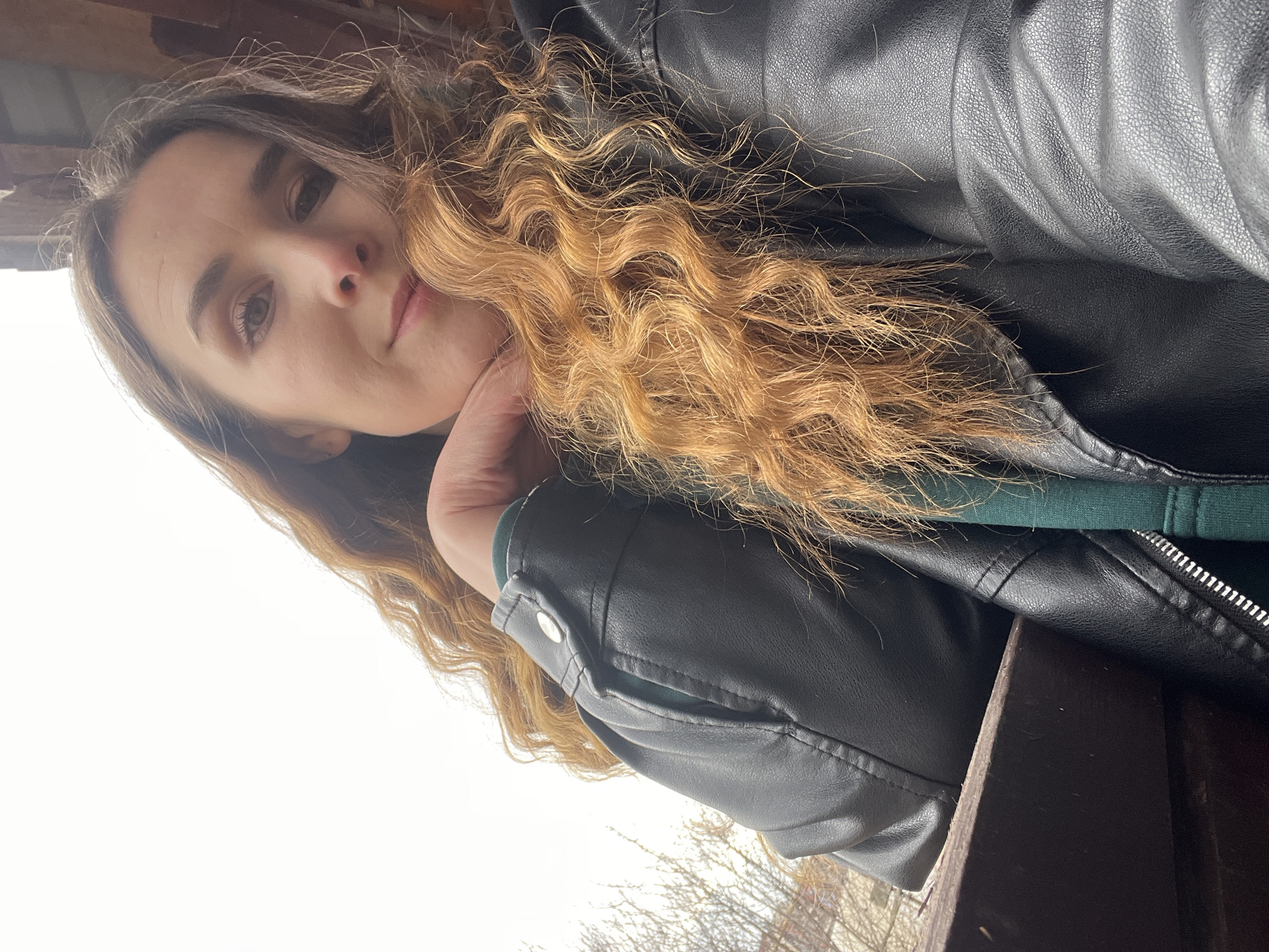 A woman is seen in an outdoor setting. The woman has long, wavy hair, brown eyes, and is wearing a black leather jacket over what appears to be a green hooded sweatshirt. She is resting her chin on her hand. The background includes bare tree branches and a wooden structure. The location cannot be determined to be Votkinsk, Russia based on the image.