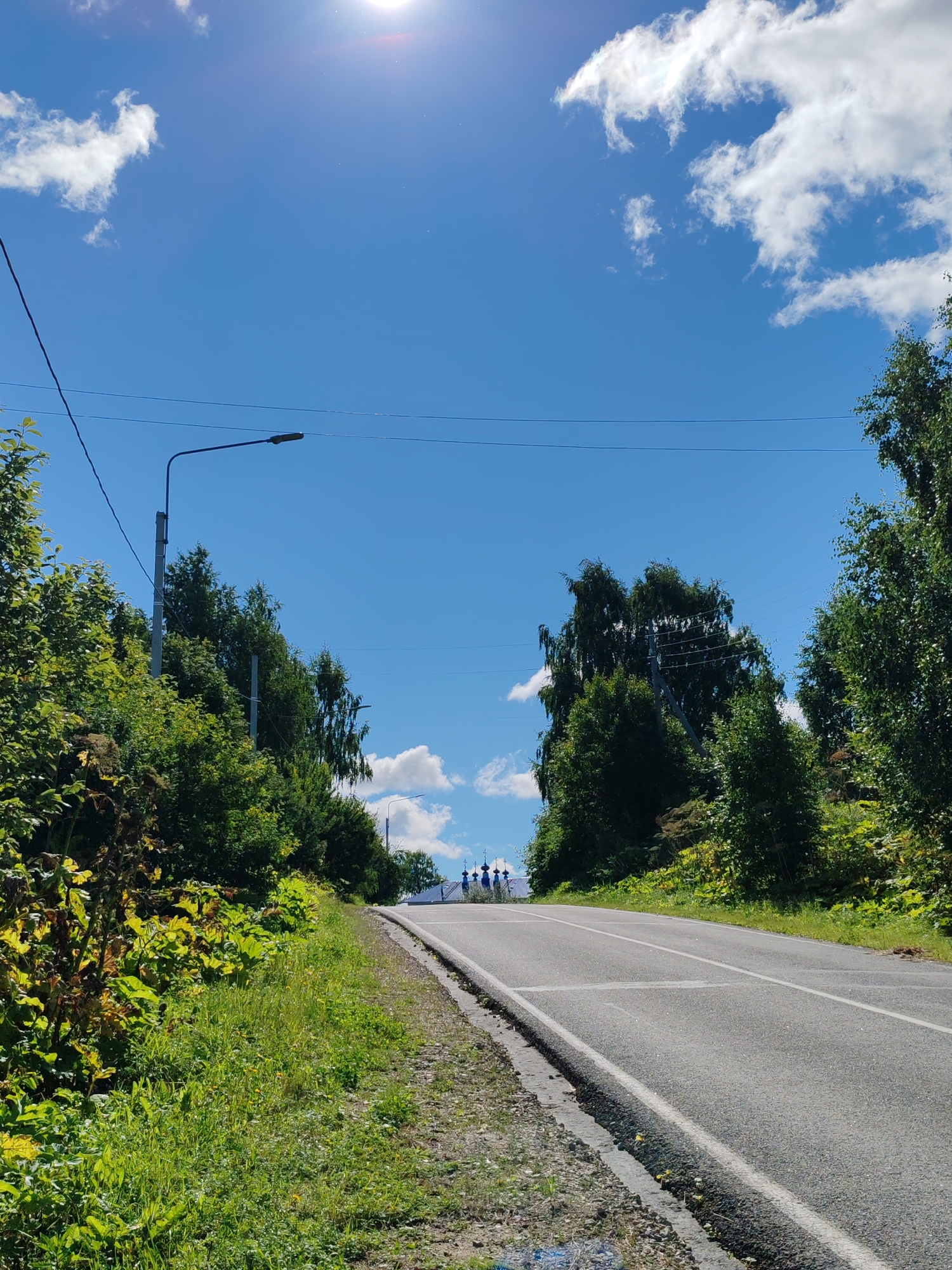 The image shows a road curving upwards through a landscape of lush green trees and vegetation under a bright blue sky with scattered white clouds. The sun is visible in the upper right corner, casting a bright glare. A lone street light stands on the left side of the road. In the distance, at the crest of the hill, several figures are visible on or near the road, and a cluster of blue-domed structures, characteristic of a Russian Orthodox church, can be seen. The scene appears to be a rural or semi-rural setting, possibly on the outskirts of the city of Chukhlem, Russia, given the presence of traditional Russian architecture. The road is paved and marked with white lines, suggesting it is a public thoroughfare. The overall impression is one of a sunny, pleasant day in a picturesque natural environment.