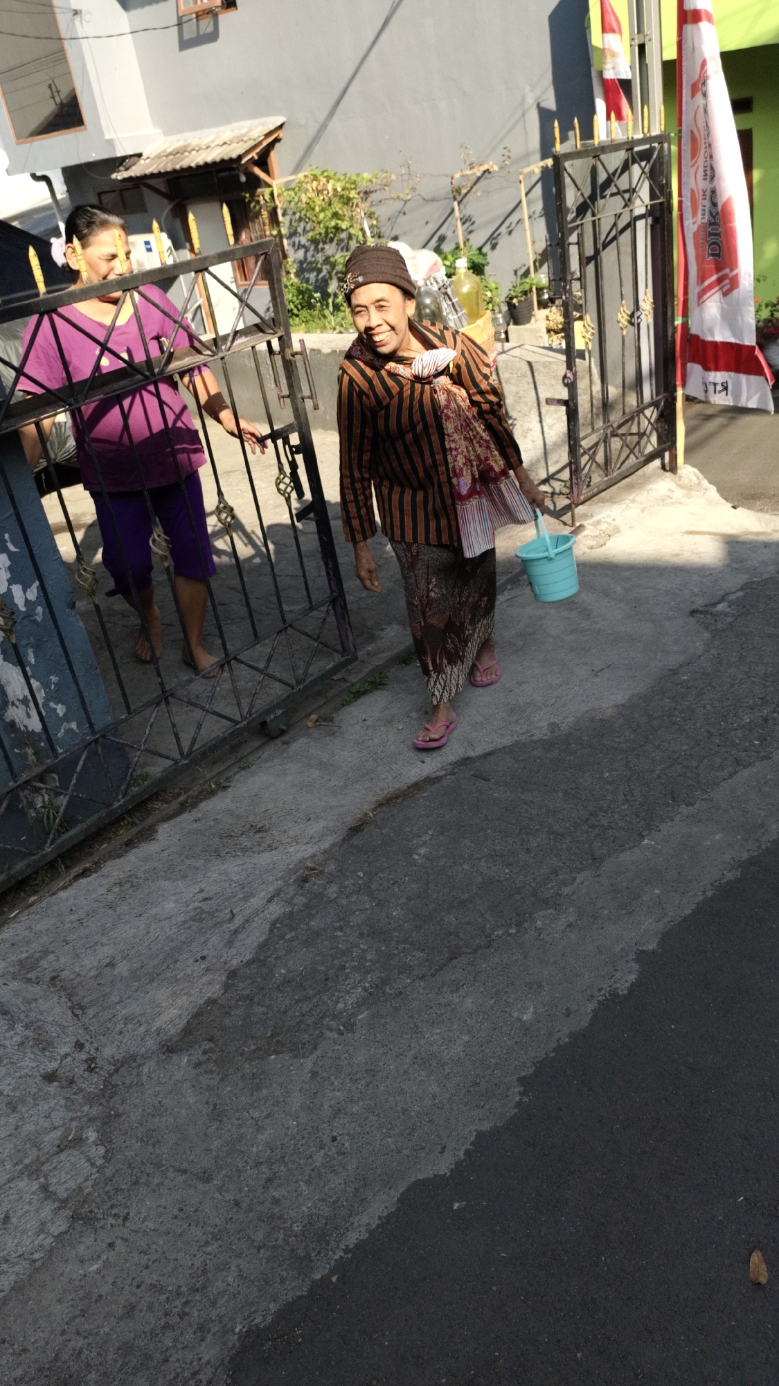 In Cimahi, Indonesia, two women are standing near a metal gate. The woman on the left, wearing a purple top and purple pants, is looking to her right. The woman on the right, who is smiling, is wearing a striped long-sleeved shirt and a patterned skirt, and is holding a light blue bucket in her right hand. She also has a patterned scarf around her neck and is wearing a brown beanie. Behind the women, a gray building is visible, with a white and red flag attached to the gate. The ground in front of them is paved with concrete and asphalt.