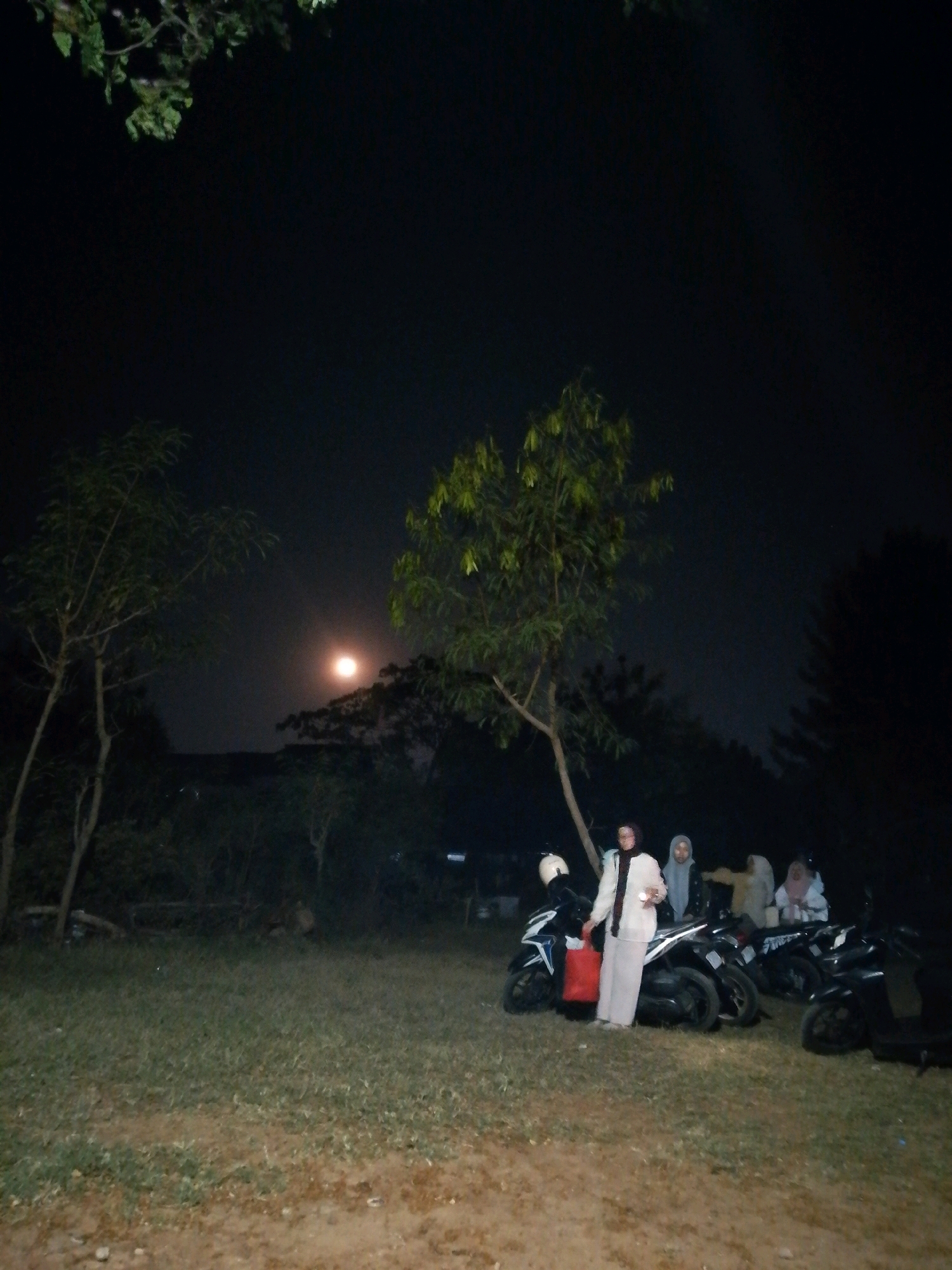 The image shows a group of people standing around several motorcycles on a grassy area at night. The sky is dark with a faint glow, suggesting a moonlit night. In the background, trees are visible, some with leaves. The lighting is dim, casting shadows and making some details indistinct. One person in the foreground is wearing a light-colored outfit and holding a red bag. Other individuals are also present, mostly wearing headscarves. The motorcycles are parked in a row. The ground appears to be a mix of grass and dirt. The overall scene suggests a casual gathering or stop during a night outing. There are no specific landmarks visible that would pinpoint the location to Cikal, Indonesia.