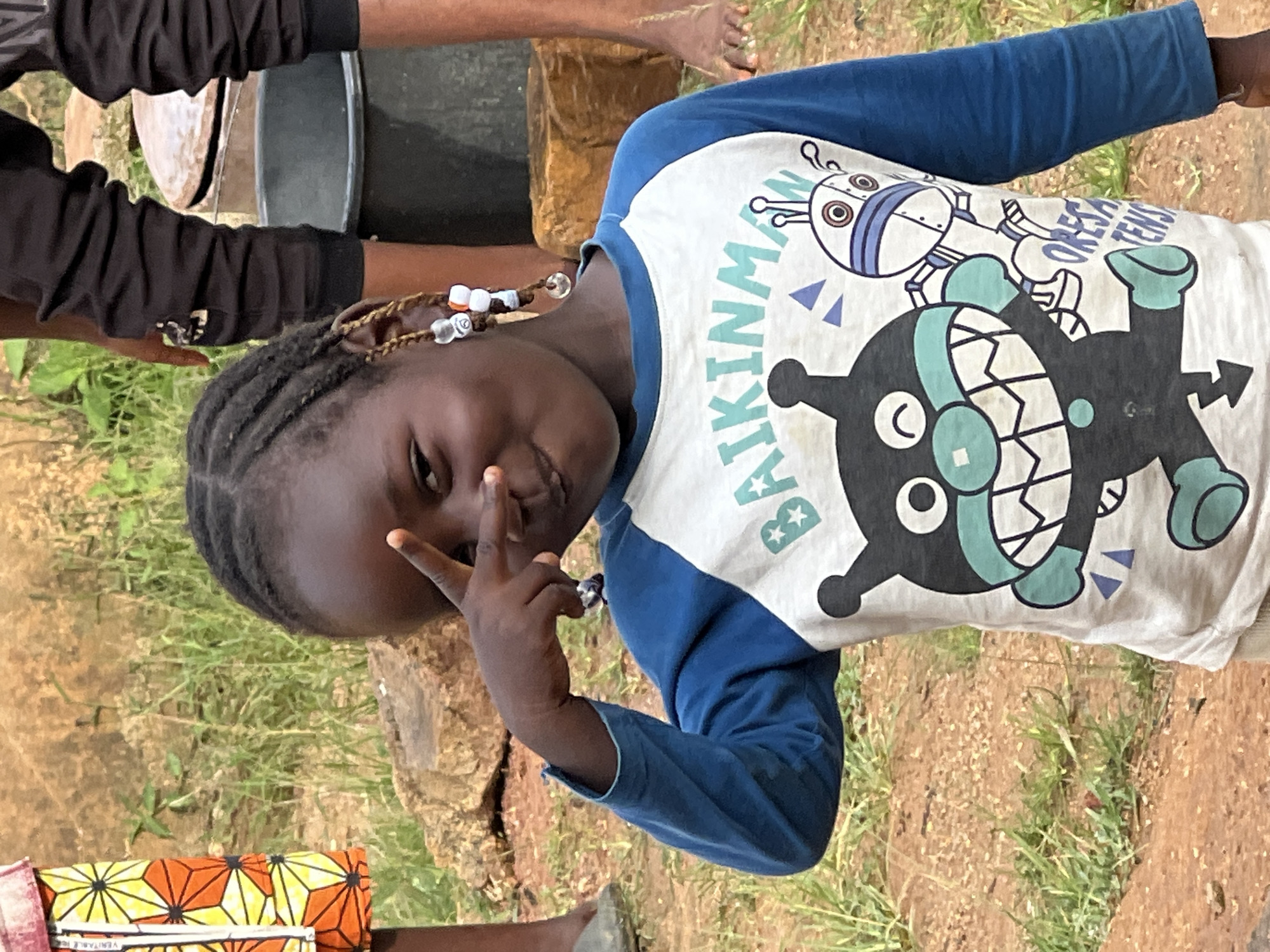 A young Black girl with braided hair adorned with beads is posing for a photograph, holding up two fingers to her forehead. She is wearing a long-sleeved shirt with a cartoon character design. The background appears to be an outdoor, rural or semi-rural setting with dirt, grass, and rocks. Other individuals' legs are visible in the background, one wearing black pants and the other wearing a colorful patterned skirt. The scene suggests a casual, everyday moment. This photograph was taken in Jos, Nigeria.