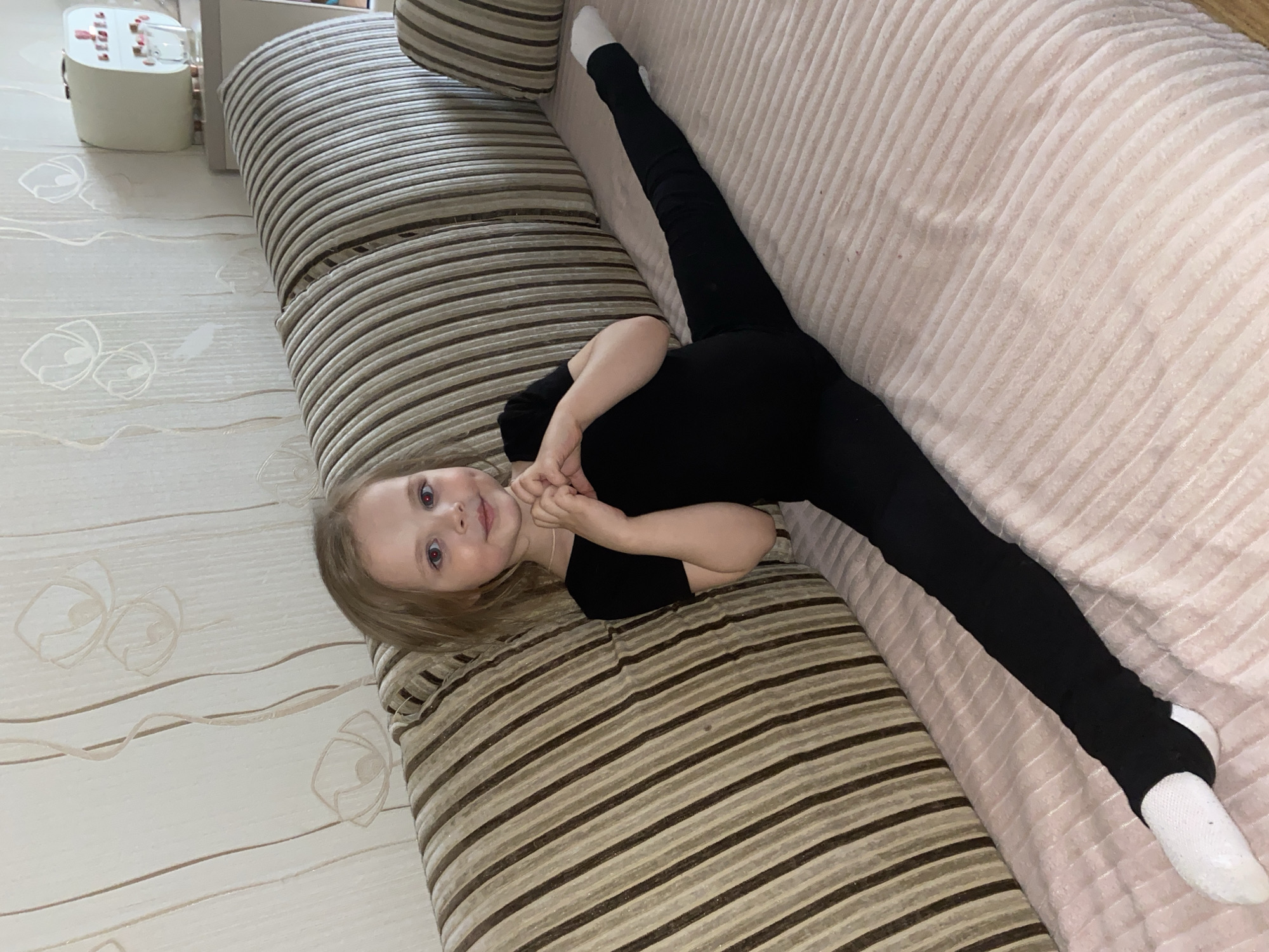 A young girl is lying on a sofa, performing a full front split or a significant leg stretch. She is positioned diagonally across the frame, with her upper body on a striped section of the sofa and her legs extended onto a ribbed, light pink/beige section. Her head is tilted towards the viewer, and she has a gentle smile, with her hands clasped together near her chest.
The girl has light brown hair and is wearing a black short-sleeved top and black leggings, which appear to be stirrup-style covering her feet, worn over white socks. Her eyes are light-colored.
The background features light-colored wallpaper with subtle floral or swirling patterns in a lighter brown or beige tone. To the left, a small, white electronic device with red buttons on top is visible on what appears to be a surface next to the sofa. The sofa itself has two distinct textures: one part is striped in varying shades of brown, and the other is a ribbed, soft pinkish-beige. The overall lighting suggests an indoor setting, likely a home in Votkinsk, Russia.