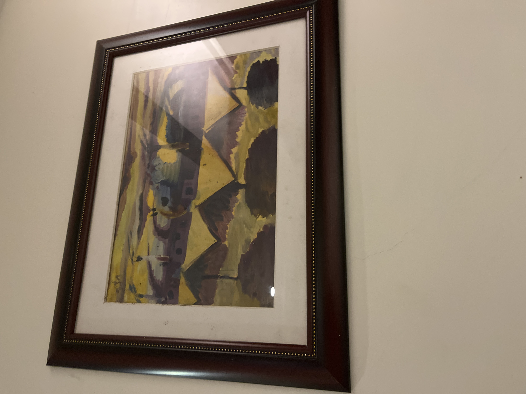 The image displays a framed abstract painting hanging on a light-colored wall. The painting itself features a composition of various geometric and organic shapes, primarily in shades of yellow, dark brown, and hints of purple, blue, and green. The style appears to be abstract, with a focus on color blocks and intersecting lines, possibly suggesting a cubist or expressionist influence.
The artwork is enclosed within a dark brown wooden frame, which has a distinct golden beaded trim along its inner edge, bordering an off-white mat. Reflections are visible on the glass covering the painting, indicating an indoor lighting source.
The wall on which the painting hangs is a light, neutral color, possibly off-white or beige. There are some faint, spiderweb-like cracks and minor scuff marks visible on the wall, especially towards the right side of the frame. The photograph is taken from a slightly low and angled perspective, looking up at the painting. There is no visible text or signature clearly discernible on the painting or the frame. The scene depicts a static interior setting, possibly in Abuja, Nigeria, as per the provided context.