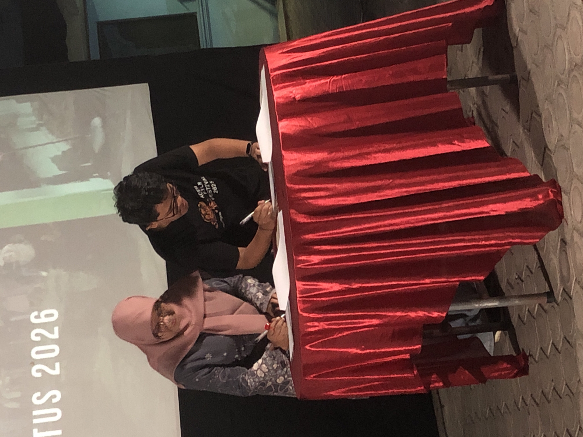 Two individuals are signing documents at a table draped with a red cloth. The person on the left, wearing a pink hijab and a blue and white patterned long-sleeved dress, is writing with a red pen. The person on the right, wearing a black t-shirt with "ACEN FILM FESTIVAL 2025" printed on it, is also writing with a pen. The background shows a large projection screen with the text "TUS 2026" visible. The scene appears to be taking place outdoors, possibly at an event or ceremony. The ground is paved with hexagonal tiles. The overall atmosphere suggests a formal event, possibly a partnership or agreement signing. This event could be related to the film festival mentioned on the t-shirt, and given the text on the screen and the general context, it is plausible that this event is taking place in Lhokseumawe, Indonesia.