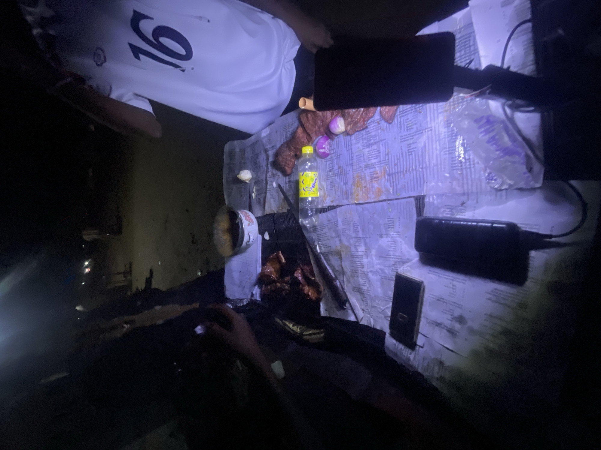 From an elevated perspective, a table with a patterned blue and white cloth sits in a dimly lit environment, possibly Lokoja, Nigeria. A person wearing a white shirt with the number "16" is visible, positioned near the top of the image. The table surface is occupied by various items: cooked meat, raw meat, onions, a yellow bottle (likely containing liquid), a knife, and other objects including a cell phone. The lighting is poor, with shadows obscuring some details. The scene shows ingredients and tools possibly for grilling or cooking. A beverage can, likely used as a cooking implement, is seen at the left. Overall, the scene presents preparations for cooking in a poorly illuminated area.
