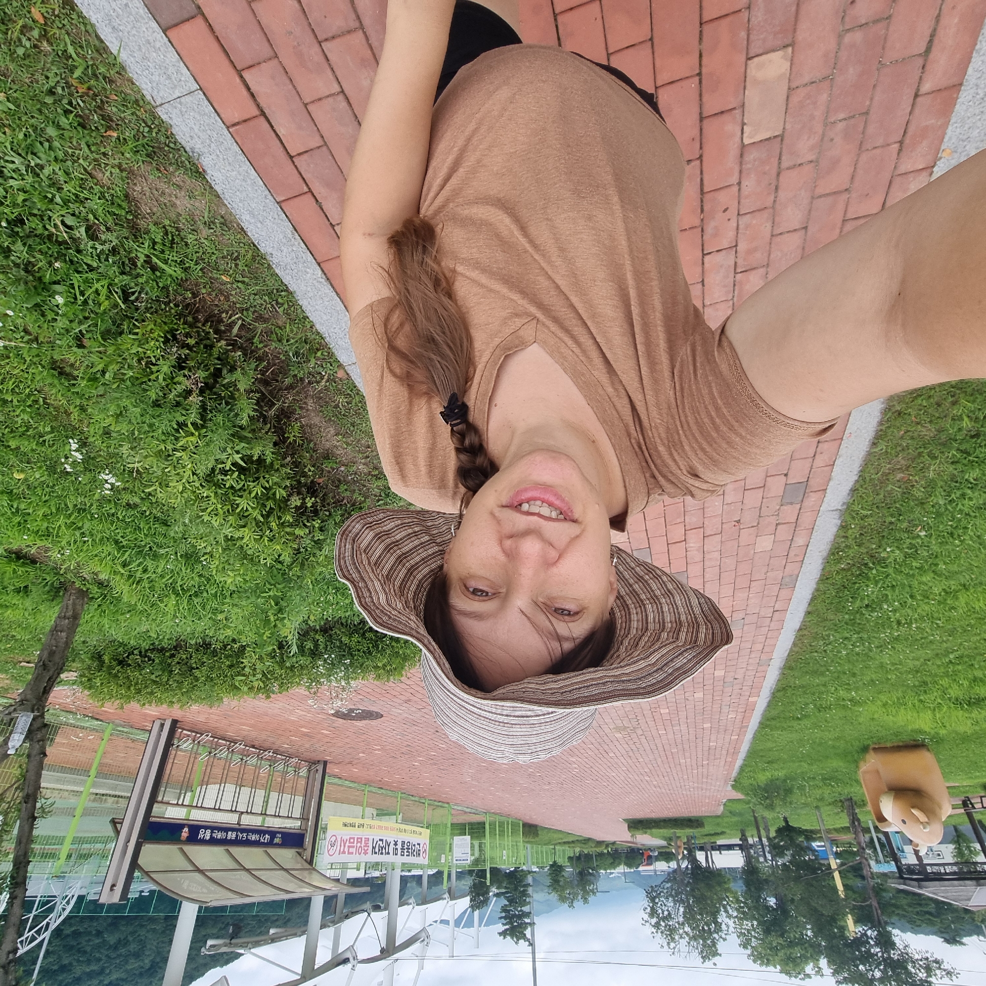 A woman wearing a wide-brimmed hat and a brown t-shirt stands on a brick pathway in Dunnae-myeon, South Korea. Her hair is in a braid. She is smiling and taking a selfie. The pathway is lined with grass and bushes. In the background, there is a fence, a bicycle rack, and a building with a white roof. Trees and a cloudy sky are visible in the distance.