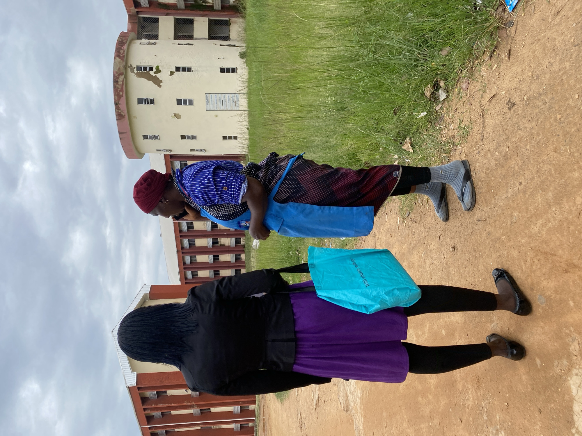 Two individuals are pictured outdoors on a cloudy day in Jos, Nigeria. The scene appears to be in front of an educational institution, with a multi-story building visible in the background. The building is cream-colored with numerous windows and some signs of wear and tear. Lush green grass grows alongside the building and a dirt path.
In the foreground, a person wearing a blue apron over a patterned garment and a red headscarf is partially visible, leaning forward. They are wearing grey socks and flip-flops. Beside them, a second person is more fully visible, facing away from the camera. They are wearing a black top, black leggings, a vibrant purple skirt, and black flat shoes. This person is holding a bright turquoise bag with text printed on it. The text on the bag reads "With wisdom, with hope".
The activity appears to be a casual encounter or a moment of rest. The cloudy weather suggests it is daytime, likely morning or afternoon. The overall impression is of an ordinary day in an urban or semi-urban environment.